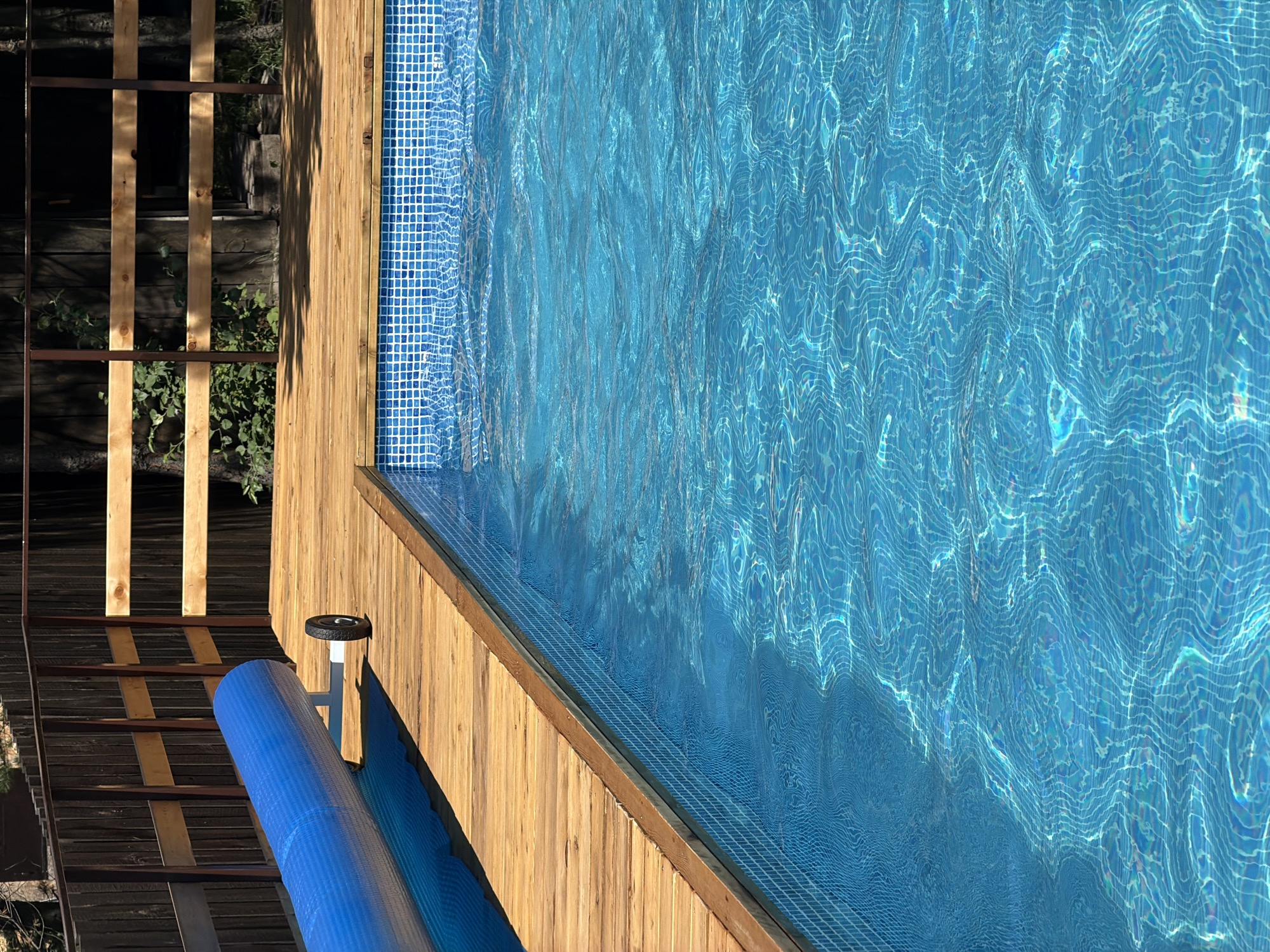 The image shows a residential swimming pool with a wooden deck. A blue pool cover is rolled up on a wheeled cart next to the pool. The pool is tiled with blue and white squares. The surrounding area includes a wooden fence and trees. The scene is located in Elekmonar, Russia.