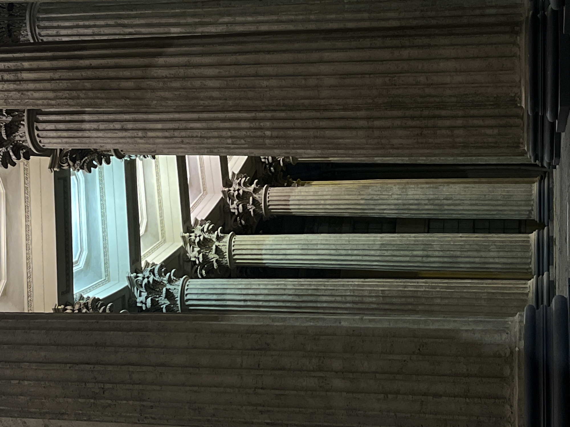 This is an upward-angled, slightly rotated, low-light photograph of classical columns in Saint Petersburg, Russia, specifically inside Saint Isaac's Cathedral. Three columns with detailed Corinthian capitals are clearly visible, with sections of columns above and below, indicating a high ceiling and multiple stories. A window is seen between the columns with an interior view of the church. The image suggests an architectural shot emphasizing the grand scale and classical design.