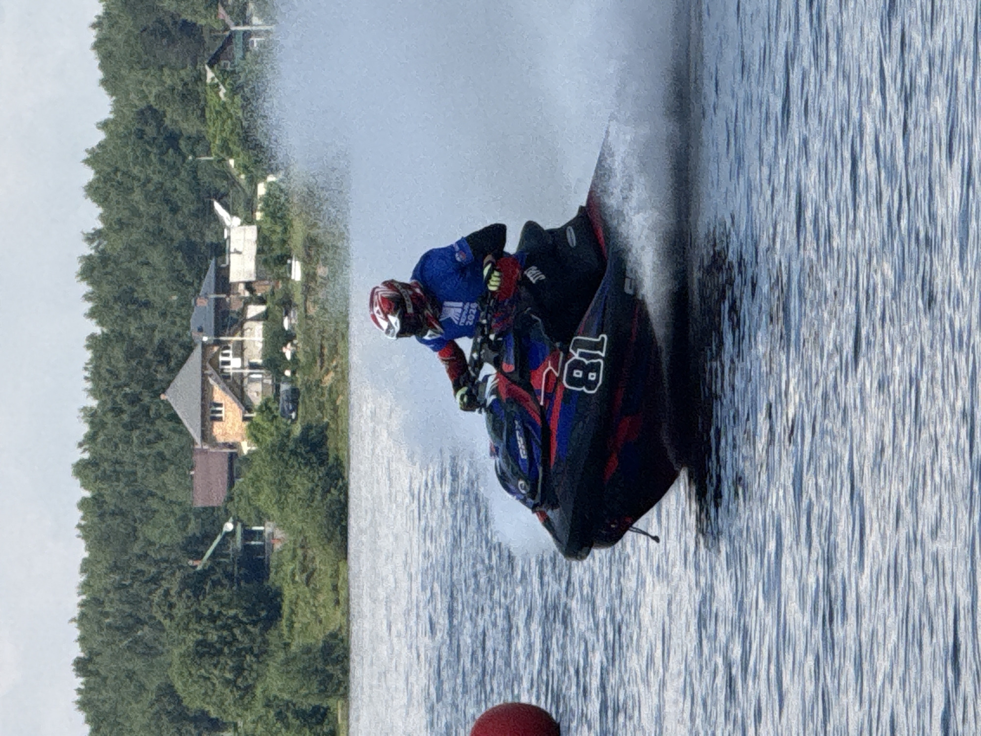 A person wearing a blue shirt with red and white accents and a red and white helmet is riding a jet ski across a body of water. The jet ski is black with red and blue accents and has the number "81" visible on its side. The rider is leaning into a turn, creating a large spray of water. In the background, a line of trees borders a shoreline with several houses. A red buoy is visible on the left side of the image. The image appears to have been taken on a cloudy day. This scene is consistent with a jet ski race taking place on the Kama River in Perm, Russia.
