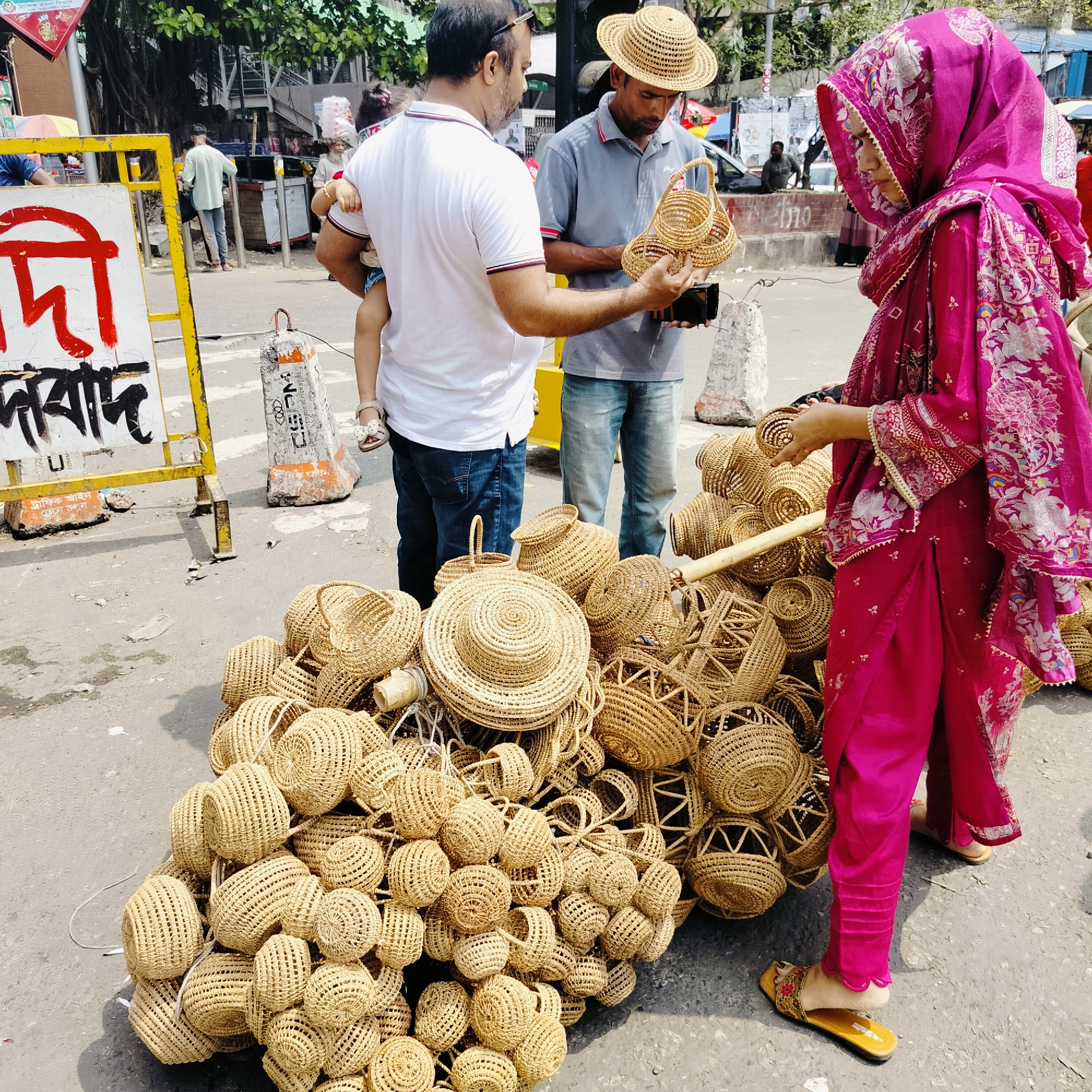 The image depicts a street scene in Dhaka, Bangladesh. A pile of woven baskets and hats occupies the foreground. A woman in a pink salwar kameez examines a basket from the pile. A man in a grey shirt and straw hat holds up smaller woven baskets for a man in a white shirt, who is holding a small child. A yellow and red sign stands to the left. The environment includes a busy street with other pedestrians and urban elements.