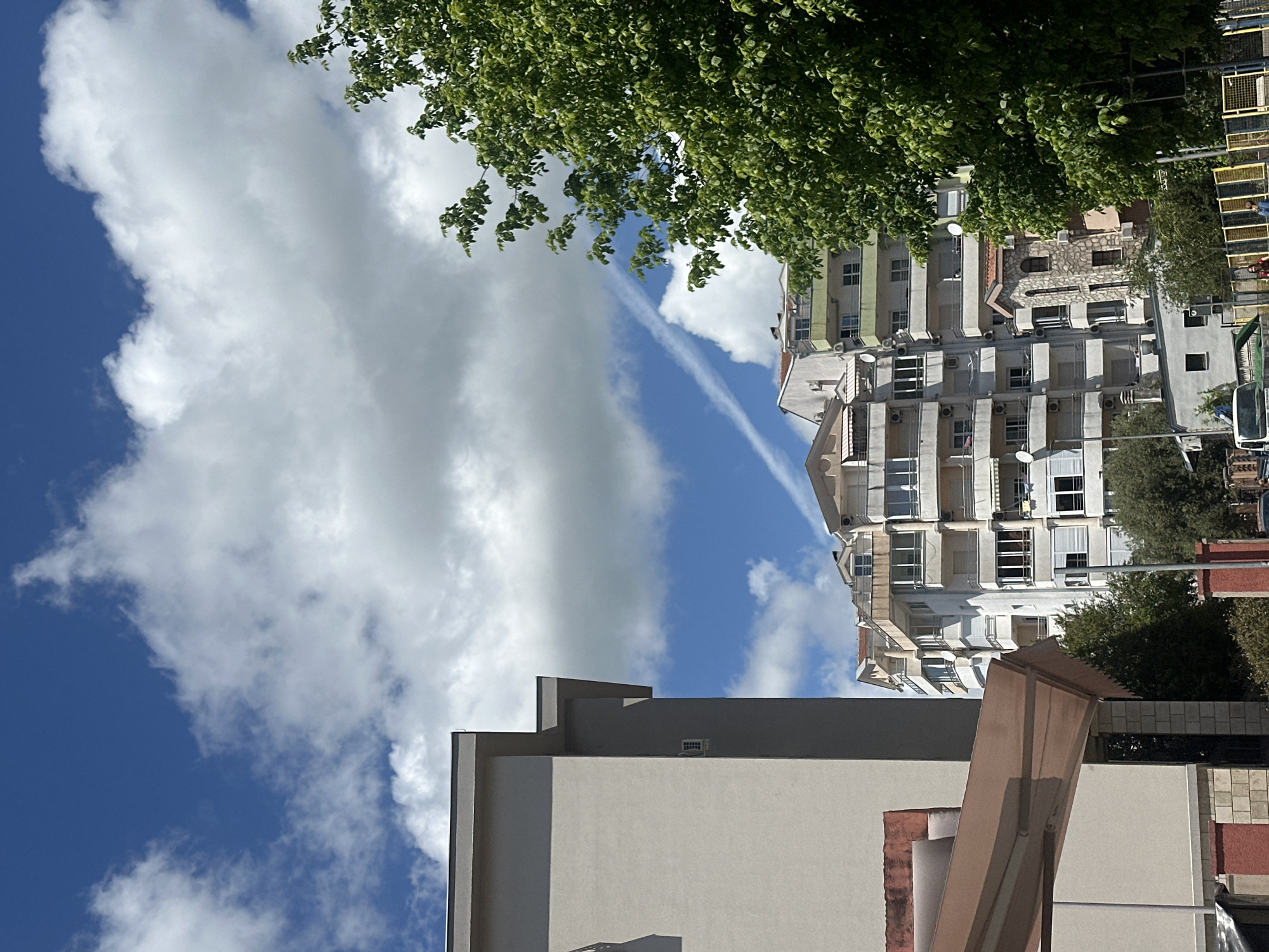The image, likely taken in Budva, Montenegro, presents a vertical view of an urban scene under a partly cloudy sky. Predominantly white cumulus clouds fill the upper left quadrant of the image, contrasting with the deep blue sky. A faint contrail stretches diagonally across the sky, cutting through the clouds.
On the right side, a multi-story residential building dominates the frame. The building features multiple balconies with white railings and numerous windows. Air conditioning units are visible on the building's facade. Dense green foliage partially obscures the building and extends to the upper right corner.
In the foreground, the corner of another building is visible. It is primarily beige with gray accents and an angular architectural element. No people are visible in the image. The overall impression is of a typical residential area within an urban setting.