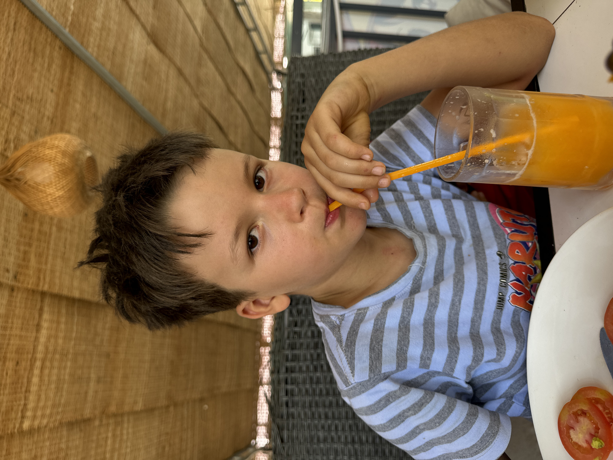 A boy with brown hair and eyes, wearing a striped blue and grey t-shirt with the words "JUMP COMICS" visible, is seated at a table, drinking orange juice from a glass with an orange straw. His left hand holds the straw to his lips. On the table, to the right of the glass, is a white plate with sliced tomatoes. The background features a woven brown wall, a grey metal pole, and a grey woven patio chair. The location is in Mũi Né, Vietnam.