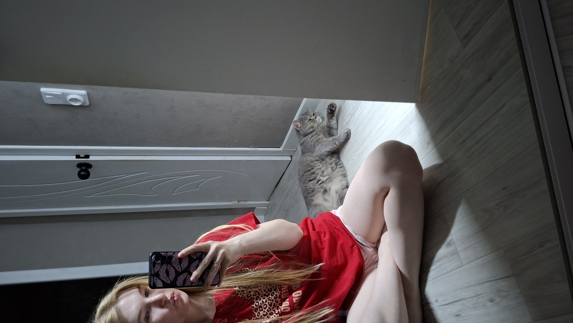 A young woman with blonde hair is taking a mirror selfie. She is sitting on a wooden floor, wearing a red t-shirt and pink shorts. She is holding a smartphone with a black and pink lip-patterned case. To her right, a grey tabby cat is lying on the floor. In the background, there is a white door and a grey wall with a light switch. The scene appears to be indoors, possibly in an apartment in Chaikovsky, Russia.