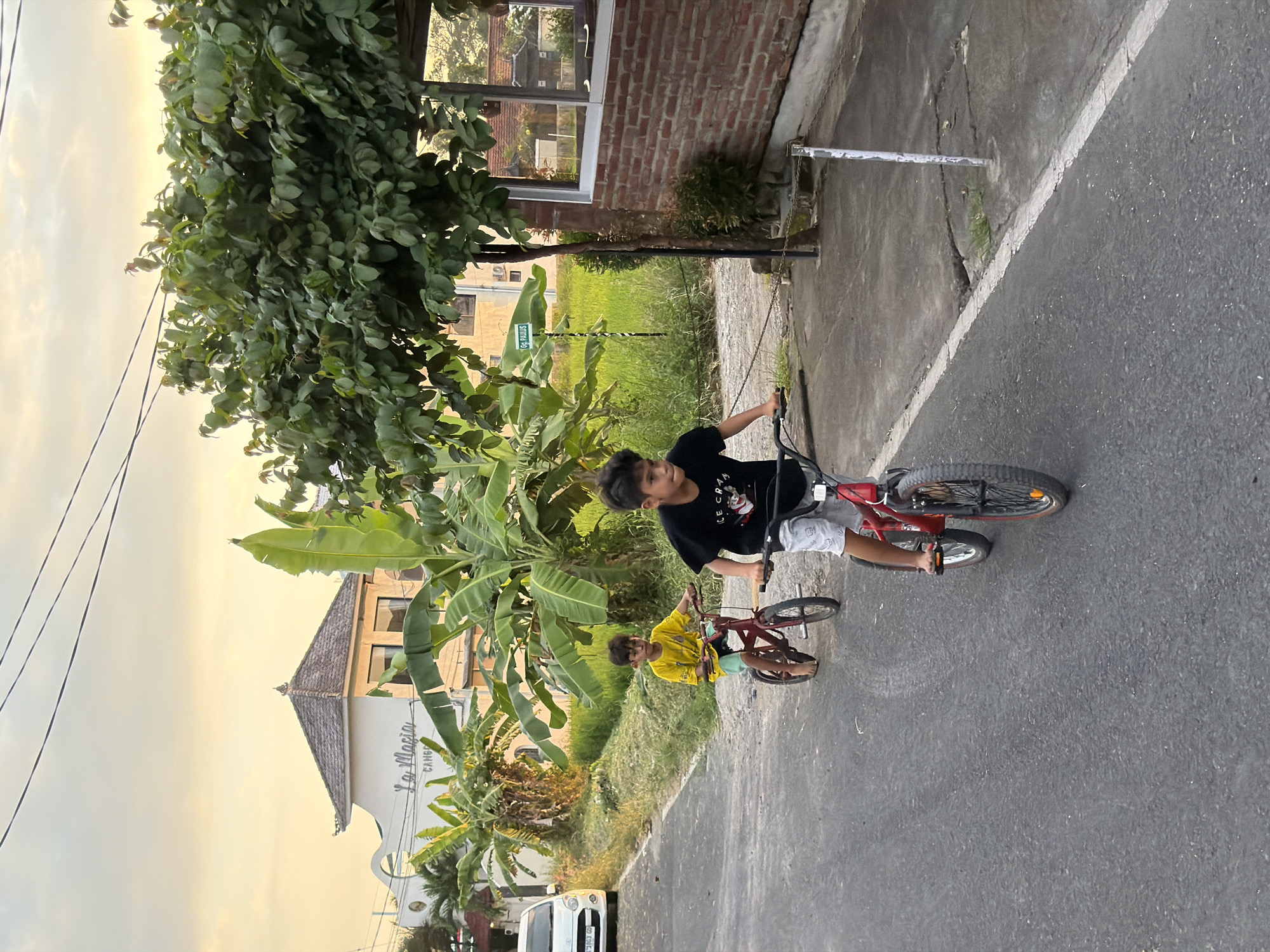 Two young boys are cycling down a paved road in North Kuta, Indonesia. The boy in the foreground, wearing a black t-shirt and light-colored shorts, is actively pedaling a red bicycle. Behind him and to his left, another boy in a yellow t-shirt and green shorts is also on a red bicycle. The road is bordered by grassy areas and vegetation, including banana trees. In the background, a white building with a pitched roof and the signage "La Macia" is visible, along with a rice paddy and a brick building. The sky is overcast with a warm, sunset glow. A white car is parked on the left side of the road. The scene appears to capture a moment of everyday life in a tropical, semi-urban environment.
