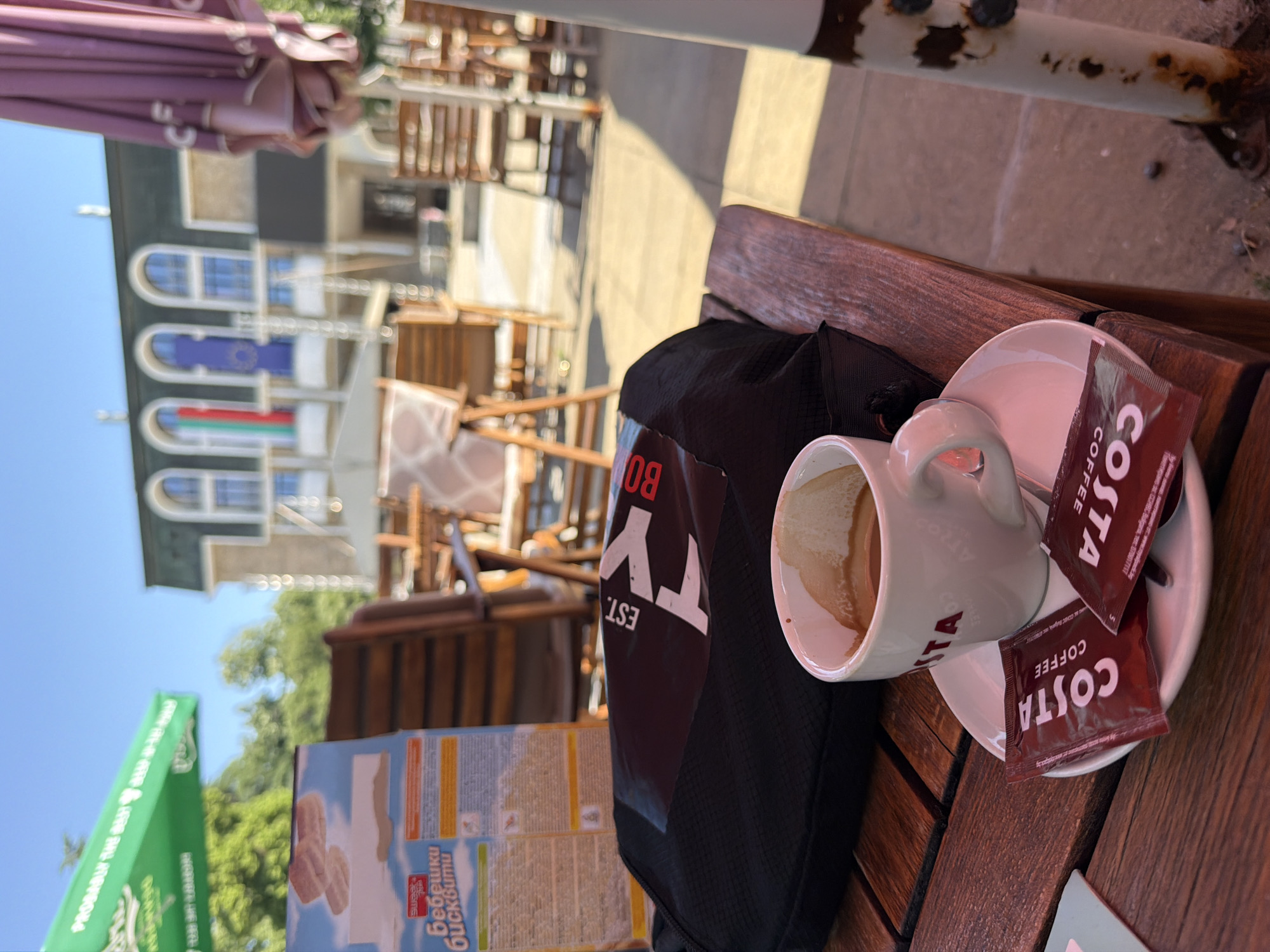 The image shows a table at an outdoor cafe in Blagoevgrad, Bulgaria. On the table is a cup of coffee from Costa Coffee, with two packets of sugar. Next to the coffee is a black bag with "BO" and "TY" printed on it. A box of Swiss cake, "бебешки бисквити", is visible. In the background are buildings, one adorned with the Bulgarian and European Union flags, and outdoor seating under umbrellas. The sky is blue.