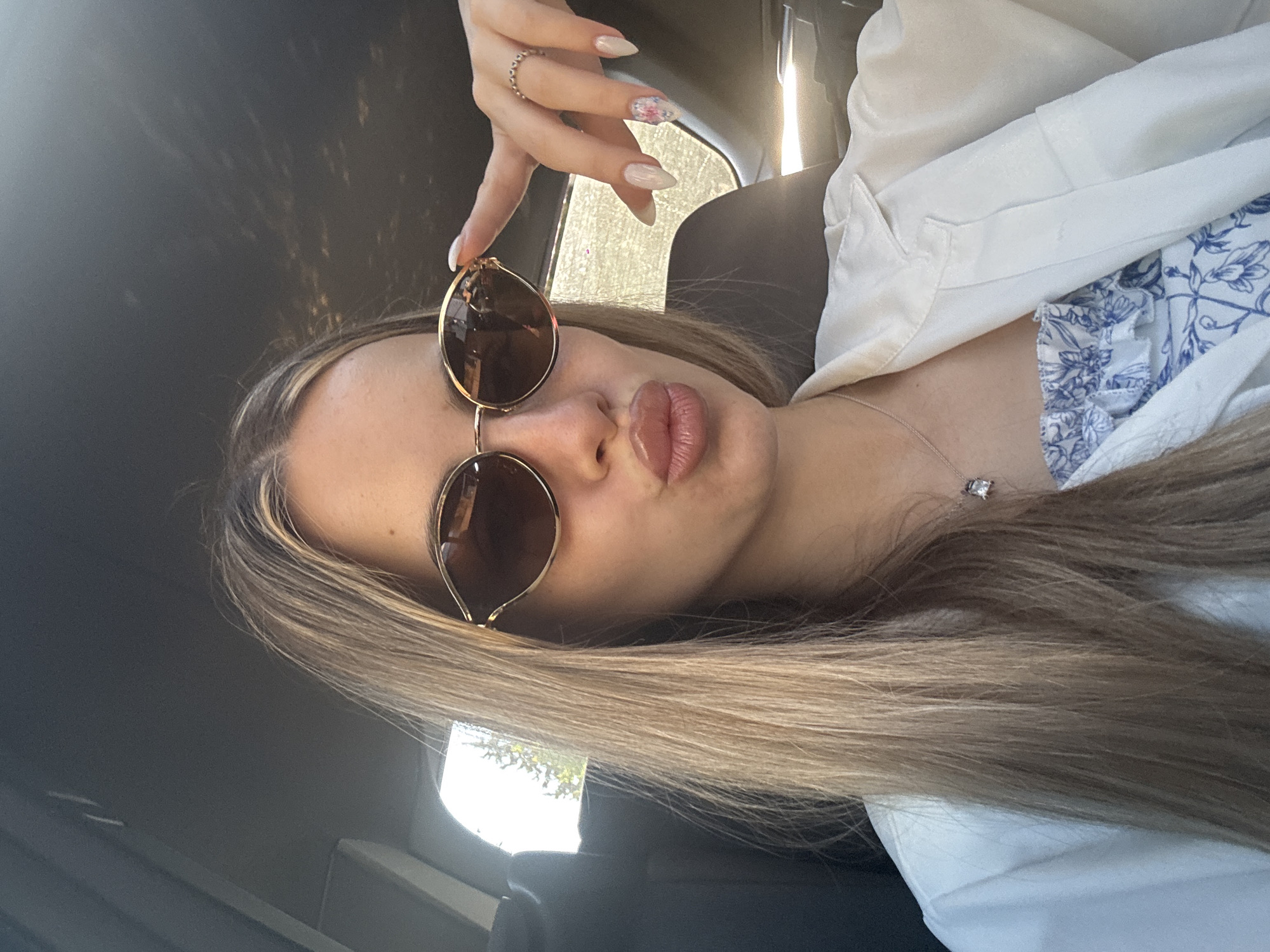In Ilindentsi, Bulgaria, a young woman with long blonde hair, wearing sunglasses, a white collared shirt, and a blue and white floral strapless top, poses for a selfie in a car. Her lips are pursed, and her right hand is raised, with her index finger touching the brim of her sunglasses. Her nails are manicured with a pearlescent finish and floral accents. A delicate necklace with a pendant is visible around her neck. The interior of the car, including the headliner and a car seat, is visible in the background, along with sunlight streaming through a window.