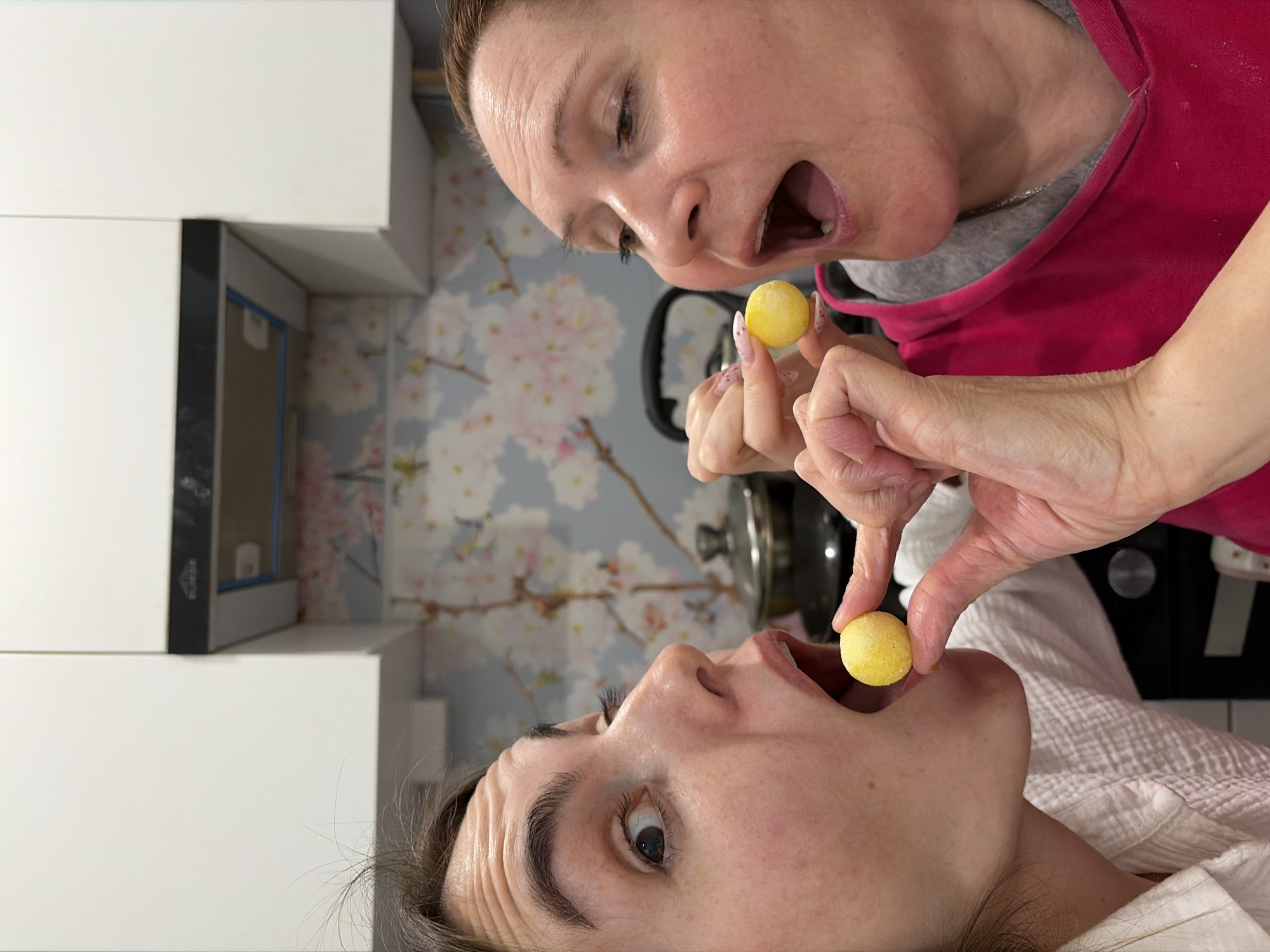 Two women are indoors in a kitchen, likely within an apartment, in Neftekamsk, Russia. One woman in a red sweater is holding a yellow ball close to the open mouth of the other, who is lying down. The kitchen has white cabinets and wallpaper with a floral pattern. A kettle is visible in the background, indicating the kitchen setting.