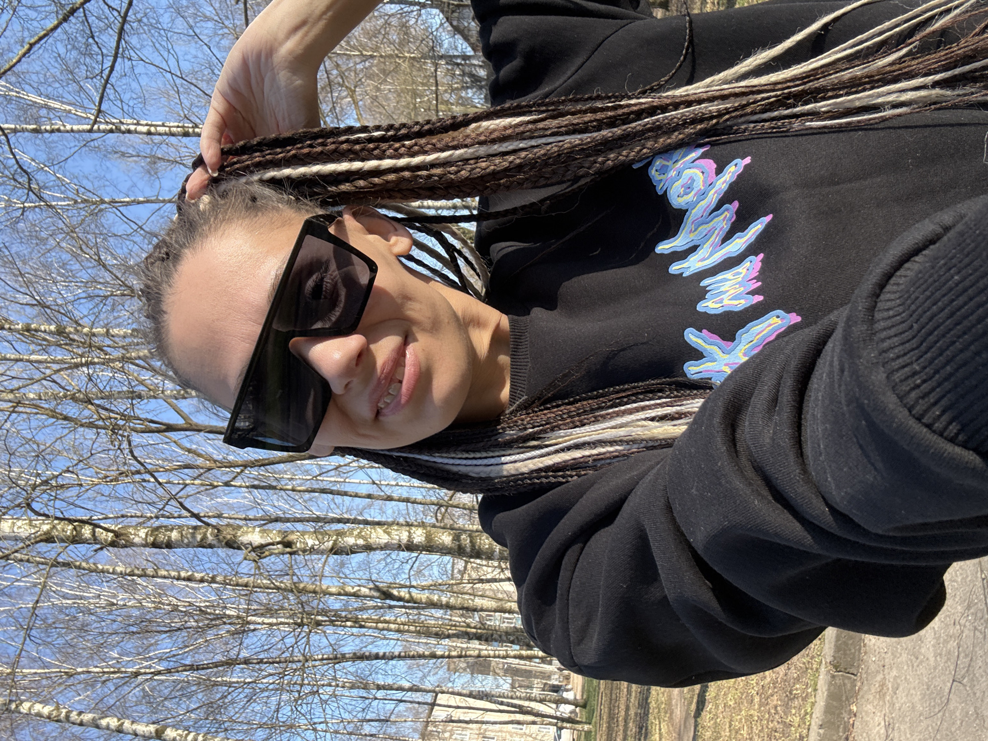 A woman with braids and sunglasses poses for a photo outside in Rusanovo, Russia. She is wearing a black sweatshirt with a colorful graphic design. Bare trees are visible in the background under a blue sky. A paved walkway is in the foreground, with a building visible further away.