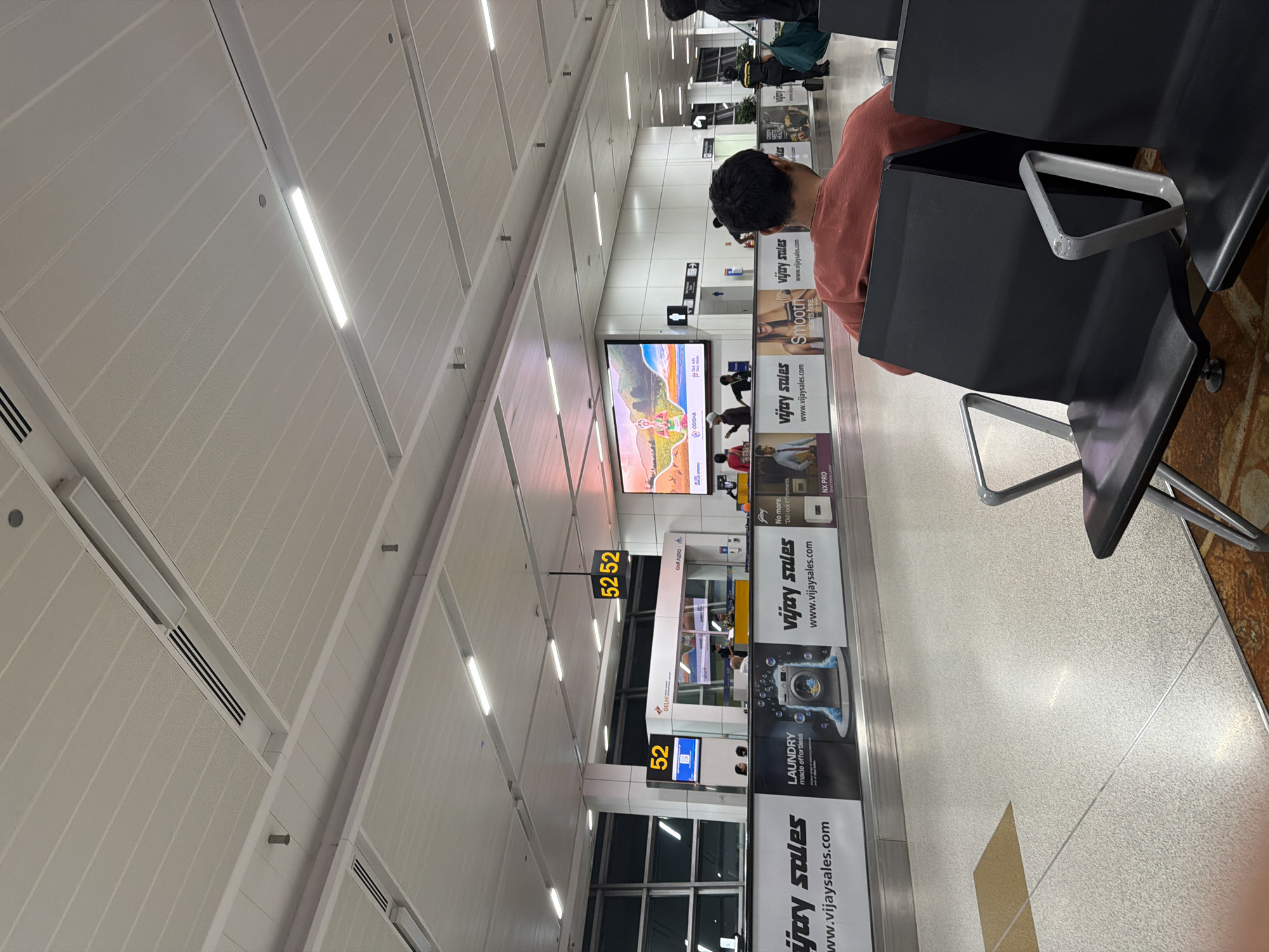 The image depicts an interior view of an airport terminal in Delhi, India. The scene is characterized by modern architecture with a high, white, slatted ceiling featuring integrated lighting and air conditioning vents. In the foreground, several rows of dark, modern airport seating are visible, with a man in a rust-colored shirt occupying one of the chairs, facing away from the camera.
In the mid-ground, a counter area with staff members is set up, suggesting a service or information point. Above this, a large digital display shows the number "52" and "52 52," likely indicating gate numbers or flight information. Adjacent to this, another large screen displays a vibrant graphic featuring a traditional Indian depiction of a woman with a golden headdress and colorful scenery. Advertising banners for "Vijay Sales" are prominently displayed along the bottom of the mid-ground, featuring slogans like "LAUNDRY made effortless" and "Smooth starts here."
The background reveals more of the terminal's architecture, including large windows, further digital screens, and directional signage. Individuals can be seen moving through the space, and security personnel in uniform are also present. The flooring appears to be a speckled light-colored material. The overall impression is one of a busy, functional airport environment.
