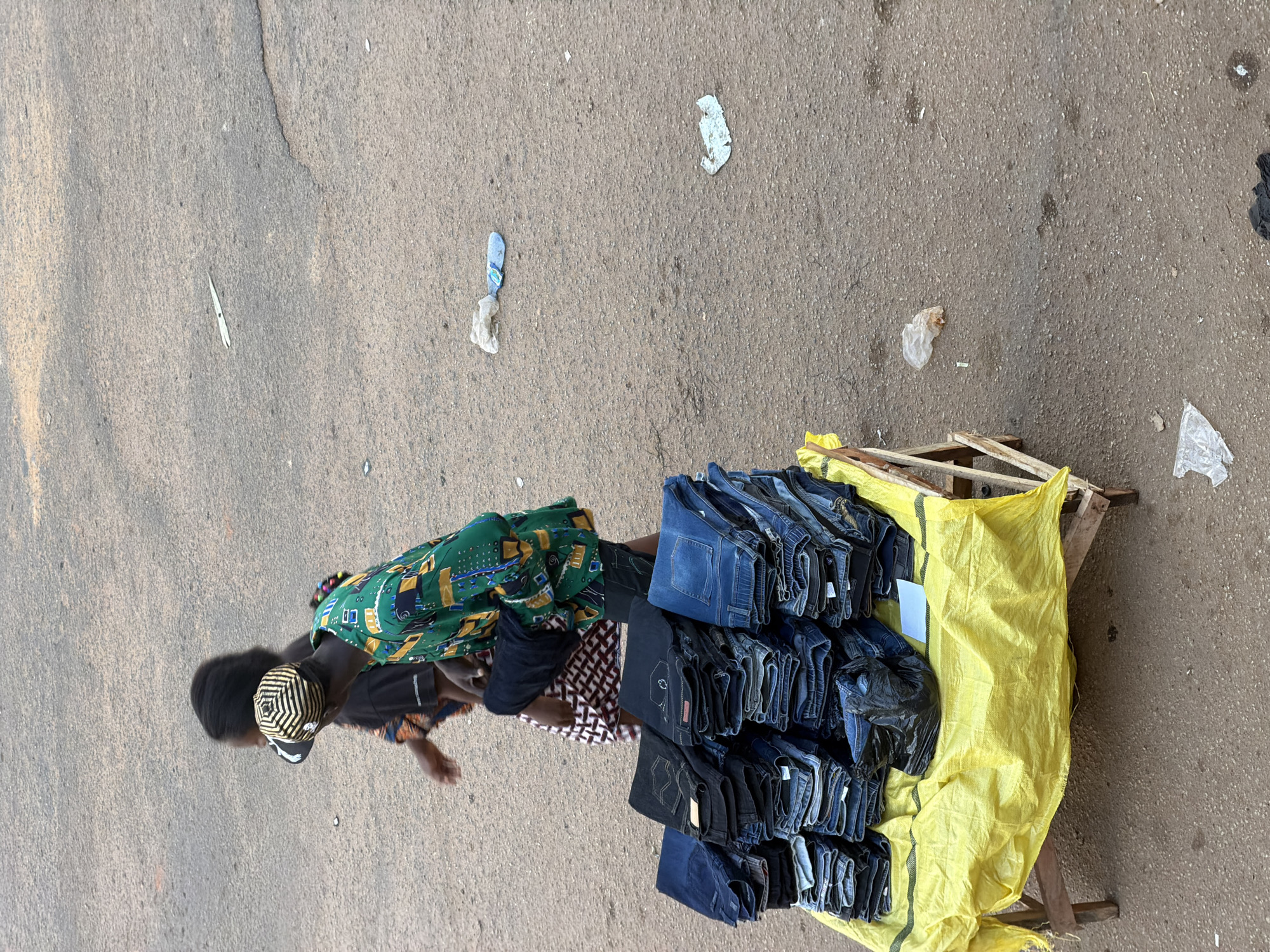 A person wearing a green patterned dress, black t-shirt, and a black and gold striped cap is seen from above in Bukuru, Nigeria. They are walking on a paved surface, possibly a street or market area. To their right, a wooden pallet covered with a bright yellow tarp holds a large stack of folded blue jeans. The jeans are neatly arranged, with many pairs visible. A black plastic bag filled with more jeans is also on the pallet, partially obscured by the yellow tarp. The ground around the pallet and the person is concrete, with scattered pieces of white and blue plastic litter. The lighting suggests it is daytime, with natural light illuminating the scene. The overall impression is of a market stall or street vendor preparing for or engaged in selling goods.