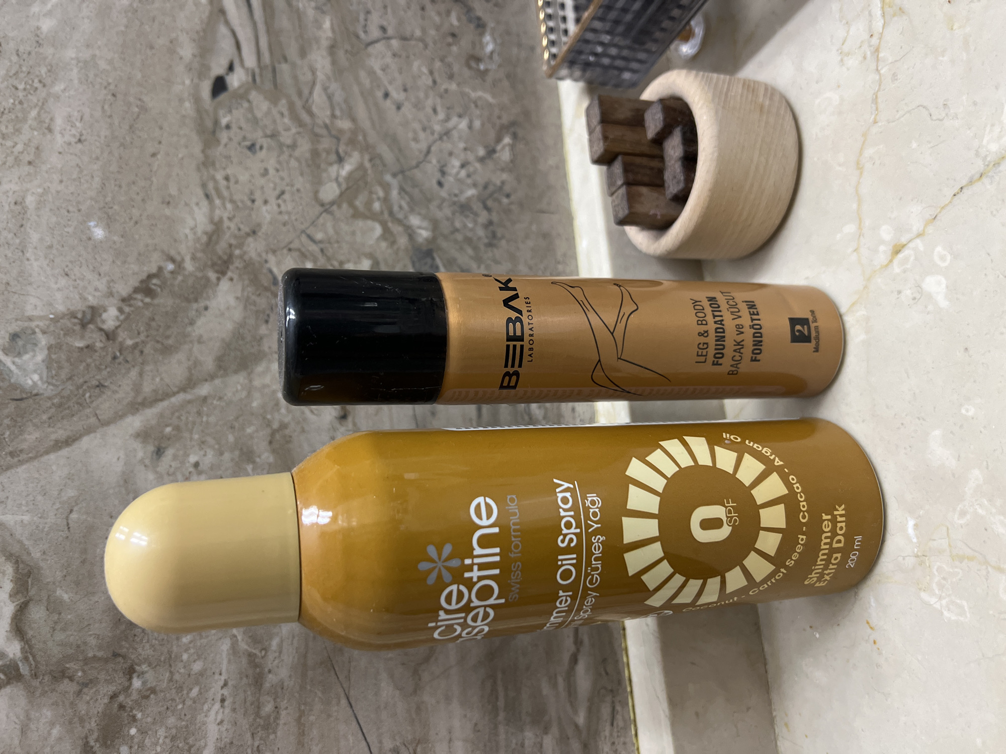 Two cosmetic products are displayed on a marble countertop in front of a marble-patterned wall. On the left is a tall, golden spray can with a light yellow cap, labeled "cire septine swiss formula shimmer oil spray". Below this, a circular sun-like graphic contains the text "0 SPF" and lists ingredients: "Carrot Seed - Cacao - Argan Oil". The bottom of the can reads "Shimmer Extra Dark 200 ml". To the right is a shorter, metallic copper spray can with a black cap, labeled "BEBAK LABORATORIES". This can features an illustration of legs and text that reads "LEG & BODY FOUNDATION Bacak ve Vücut Fondöteni", followed by "2 Medium Tone". To the right of the spray cans, a small, light-colored wooden bowl contains several dark brown wooden cubes. The scene is indoors, with no indication of a specific location within Chișinău, Moldova.