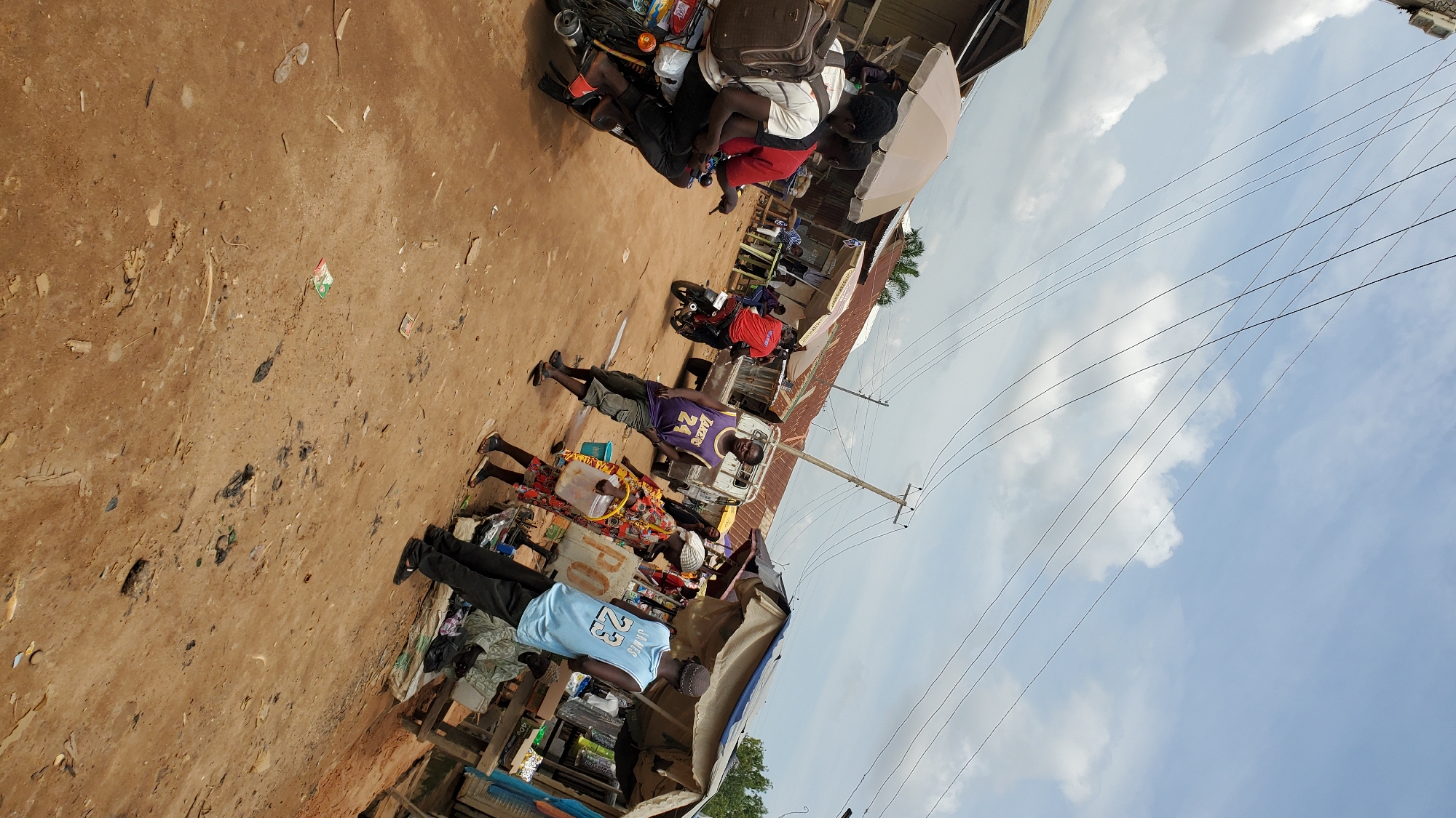 This is a photograph of a street market in Jalingo, Nigeria, under a partly cloudy sky. The ground is a dirt road. In the foreground, three young men are on a motorcycle. The rider in the back is wearing a backpack and a white t-shirt. The man in the middle is wearing a red t-shirt, and the man in the front is wearing a black t-shirt. To the right, a man in a purple tank top with the number "24" on it is walking towards the right, holding a yellow bucket. Behind him, a person in a brightly colored dress is also walking towards the right. A man in a light blue tank top with the number "23" on it is standing to the right of the person in the colorful dress. In the background, there are several market stalls with awnings, a white truck, and other people. Power lines are visible in the sky.