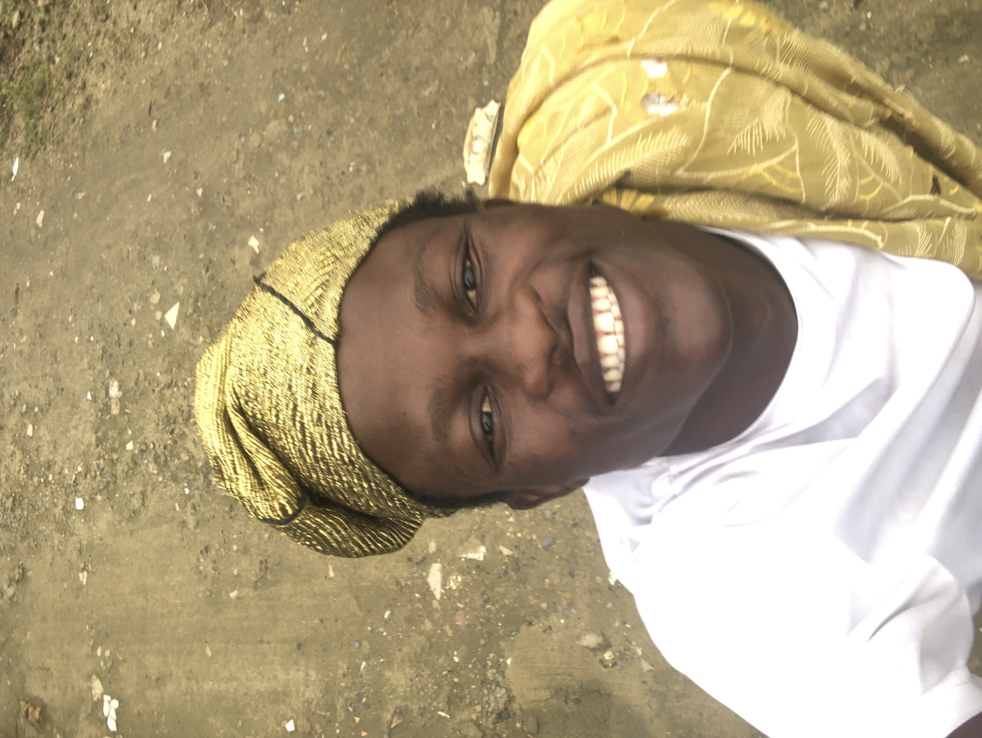 The image is a selfie taken from a high angle, looking down at a young Black male. He is wearing a white t-shirt and a gold patterned headwrap and matching garment draped over his shoulder. The man is smiling broadly, showing his teeth. The background appears to be an outdoor, unpaved area with scattered debris. The lighting suggests it is daytime. The scene is consistent with an outdoor environment in a neighborhood in Ado, Nigeria.