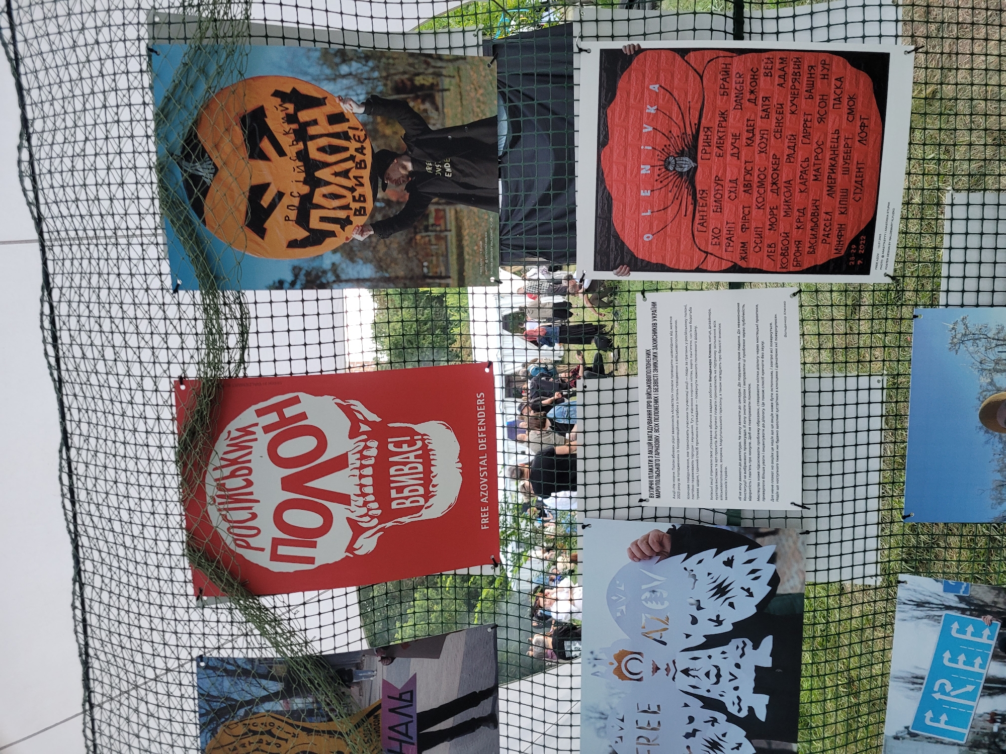 The image shows a series of posters displayed outdoors, attached to a wire mesh fence. The posters are predominantly red, white, and black, with some featuring illustrations and text in Ukrainian. One poster depicts a white skull with red text that reads "РОСІЙСЬКИЙ ПОЛОН ВБИВАЄ!" (Russian Captivity Kills!) and "FREE AZOVSTAL DEFENDERS" at the bottom. Another poster features a stylized orange pumpkin with similar text. In the background, a crowd of people is visible, suggesting an outdoor event or demonstration. The overall impression is one of protest or awareness campaign, likely related to the conflict in Ukraine. The location is identified as Unknown, Unknown.