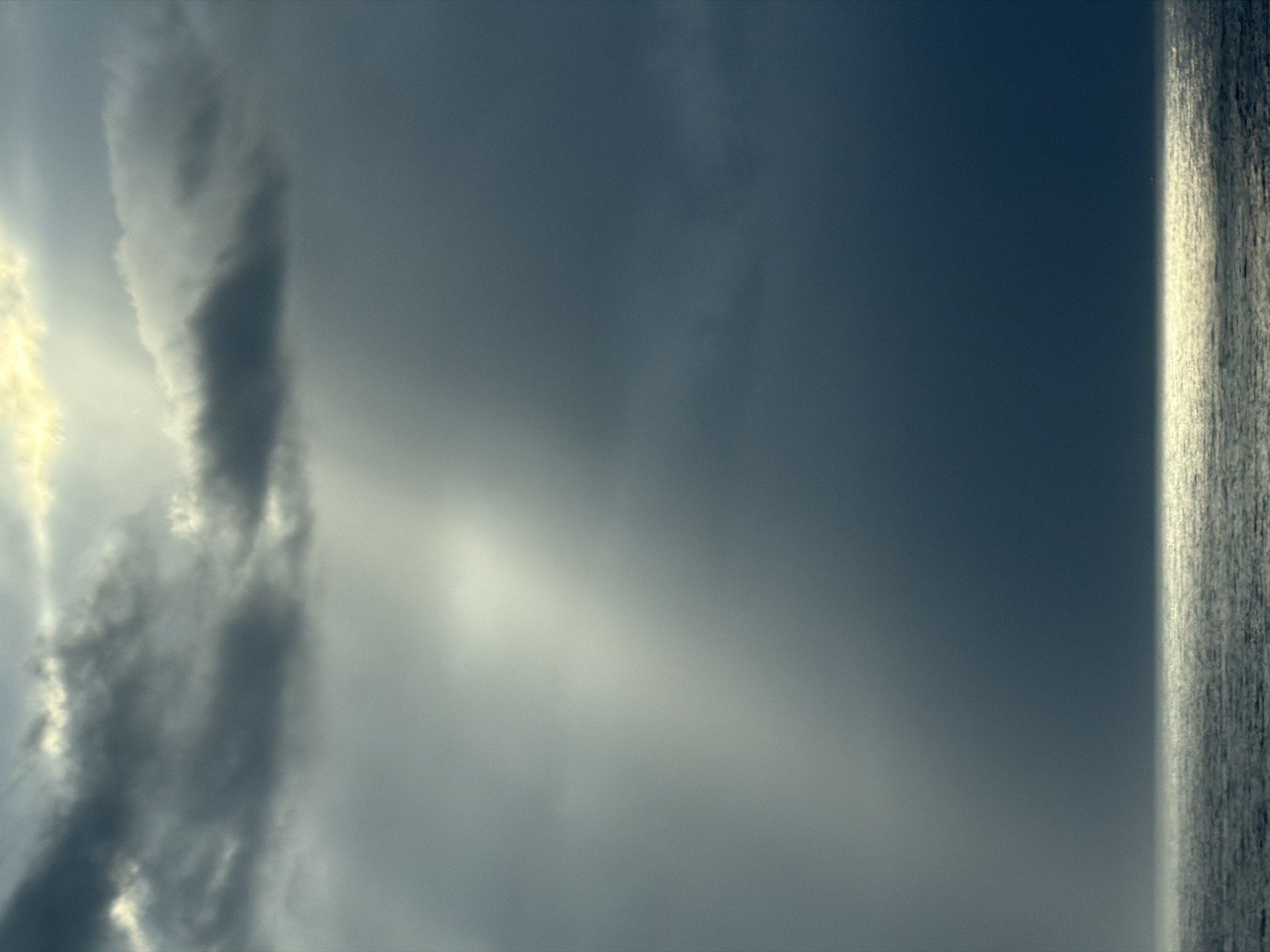 The media file displays a dramatic seascape or skyscape, best viewed when rotated 90 degrees counter-clockwise to a horizontal orientation. The majority of the frame is filled with a vast and cloudy sky. On the left side, dense, dark, and turbulent cloud formations are illuminated by bright, almost golden light breaking through from behind, suggesting the sun's presence. Rays of light are visible piercing through the cloud cover. Towards the center and right, the sky transitions to a softer, muted blue-grey, still featuring subtle cloud textures.
At the bottom of the image, a narrow, textured horizontal band represents a large body of water, likely an ocean, meeting the sky at a well-defined horizon line. The water's surface exhibits subtle ripples or reflections, and its color generally aligns with the darker tones of the sky, with a slight luminosity along the horizon.
The interplay of bright light and heavy clouds suggests dynamic weather conditions, possibly after a storm, or atmospheric light characteristic of dawn or dusk. There are no people, man-made objects, or visible text within the image. The scene conveys a sense of natural power and expansive tranquility.