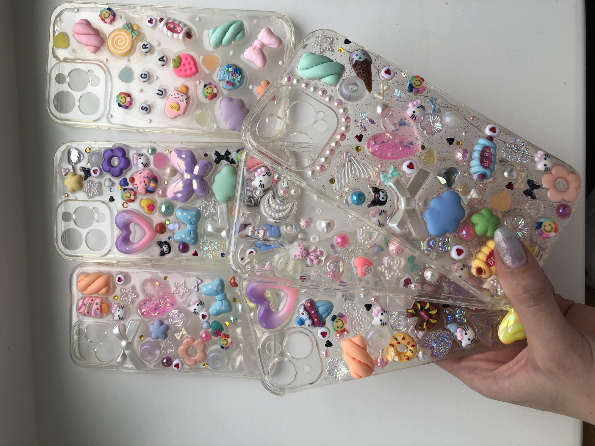 The image shows four decorated transparent phone cases stacked on a white surface, held by a woman's hand. The cases are adorned with various small decorative items, including miniature sweets, bows, hearts, flowers, and character figures. The decorations are colorful and give a whimsical, childlike appearance. The hand holding the phone cases has a fingernail painted with a glittery polish. There is no information in the picture about the location or city of Noviy, Russia.