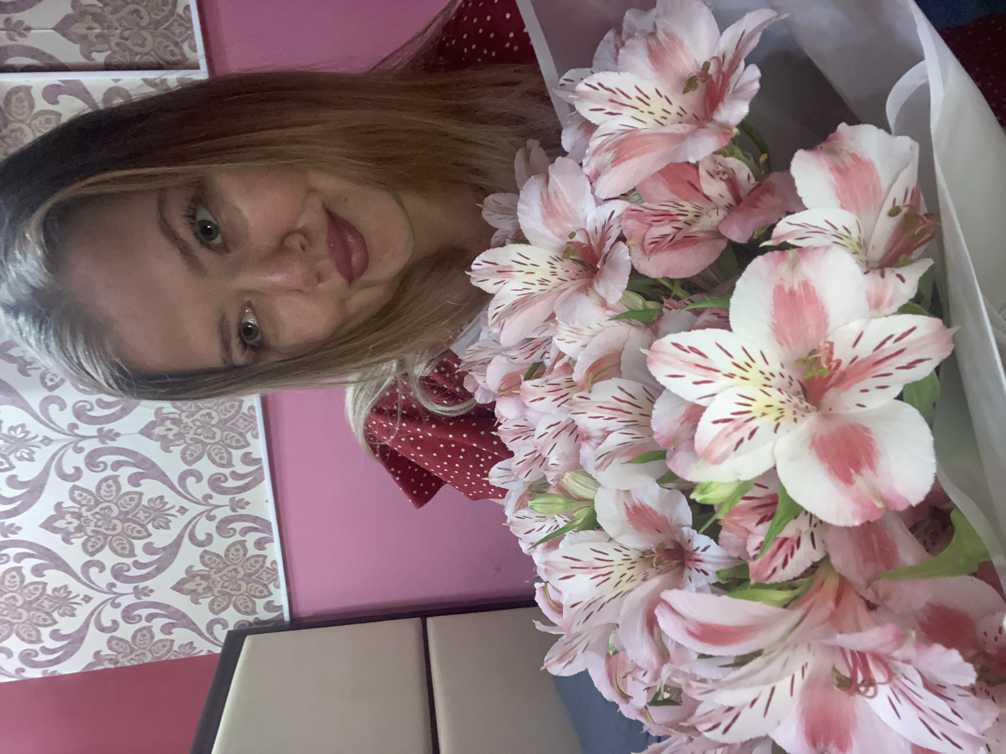 A medium shot captures a young woman holding a bouquet of pink and white alstroemeria flowers. She is positioned in the foreground, looking slightly to her right with a subtle smile. The flowers, in various stages of bloom, fill the lower portion of the frame. Behind her, a patterned wallpaper in shades of cream and mauve adorns the wall. To the left, a portion of a wooden headboard is visible, and the wall behind it is painted pink. The woman is wearing a red polka-dot dress. The overall scene suggests an indoor setting, possibly a bedroom. The image does not contain any information that would allow for its location to be identified in Yerevan, Armenia.