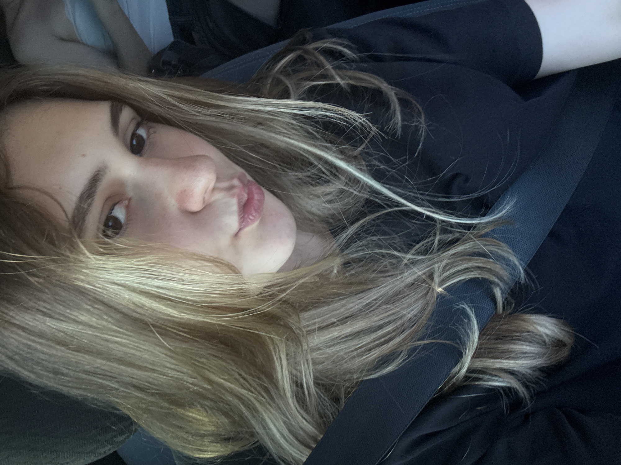 This is a close-up, selfie-style photograph taken from a slightly upward angle of a young person with blonde hair, possibly in a car, as indicated by a seatbelt visible across their chest. The person is looking towards the camera, with their head tilted. Their features are soft, and their lips are slightly parted, creating a pouty expression. The lighting suggests it's daytime, but the ambient light is somewhat subdued, possibly indicating an overcast day or indoor setting. The background is out of focus, but hints of dark fabric and possibly a headrest are visible, reinforcing the idea of being in a vehicle. The person's hair is a prominent feature, long and wavy, with streaks of lighter blonde. There is no visible text in the image. The overall mood is casual and introspective. Without further context, it is difficult to determine the specific activity or event taking place beyond a personal moment captured for a photo. The location context provided, Koprivshtitsa, Bulgaria, does not offer any visual cues within the image itself.