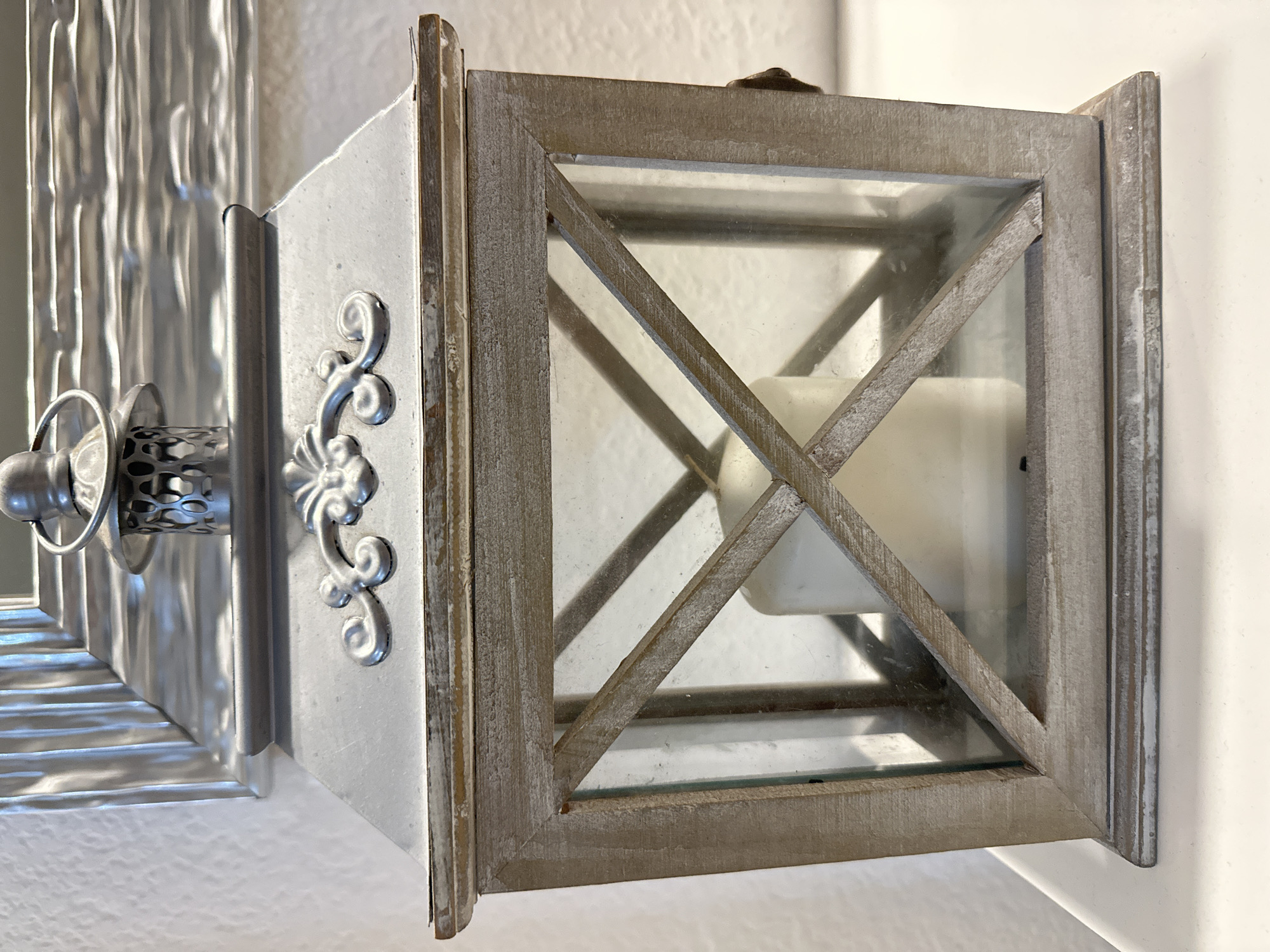 The image shows a wall-mounted decorative lantern. The lantern is primarily composed of a weathered-looking light grey wood frame, with glass panels enclosing the sides. Inside the lantern is a white pillar candle. The mounting fixture is silver with decorative flourishes and a knob-like handle. The wall behind the lantern is plain and off-white. The location within Sant Feliu de Guíxols, Spain is indeterminable from the image.