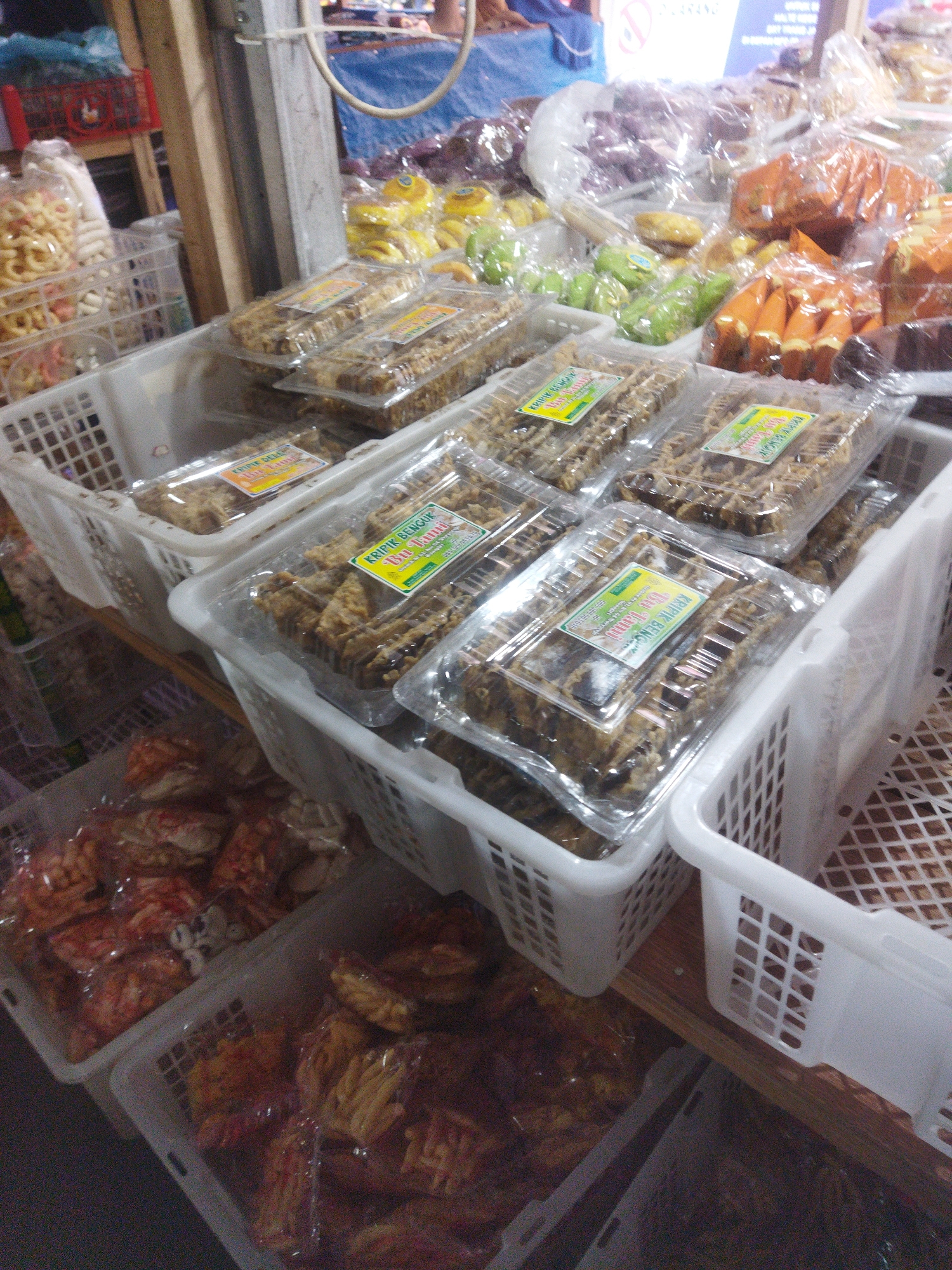 This is a close-up, eye-level shot of a market stall in Wonogiri, Indonesia, featuring an array of packaged snacks. The primary focus is on several white plastic baskets filled with various bagged and boxed food items. In the foreground, two white baskets hold numerous rectangular clear plastic containers filled with what appears to be fried snacks, likely "kripik" (chips). These containers are stacked and some have yellow and green labels indicating the product name and brand. To the left, a basket contains bags of crunchy, light-colored snacks. Further back, other baskets are visible, some holding green and orange colored snacks in plastic packaging, and some with larger bags of snacks. The stall is set up on a wooden surface. The background is blurred but suggests a bustling market environment with other stalls and people. The lighting is somewhat dim, contributing to a slightly grainy appearance of the image.