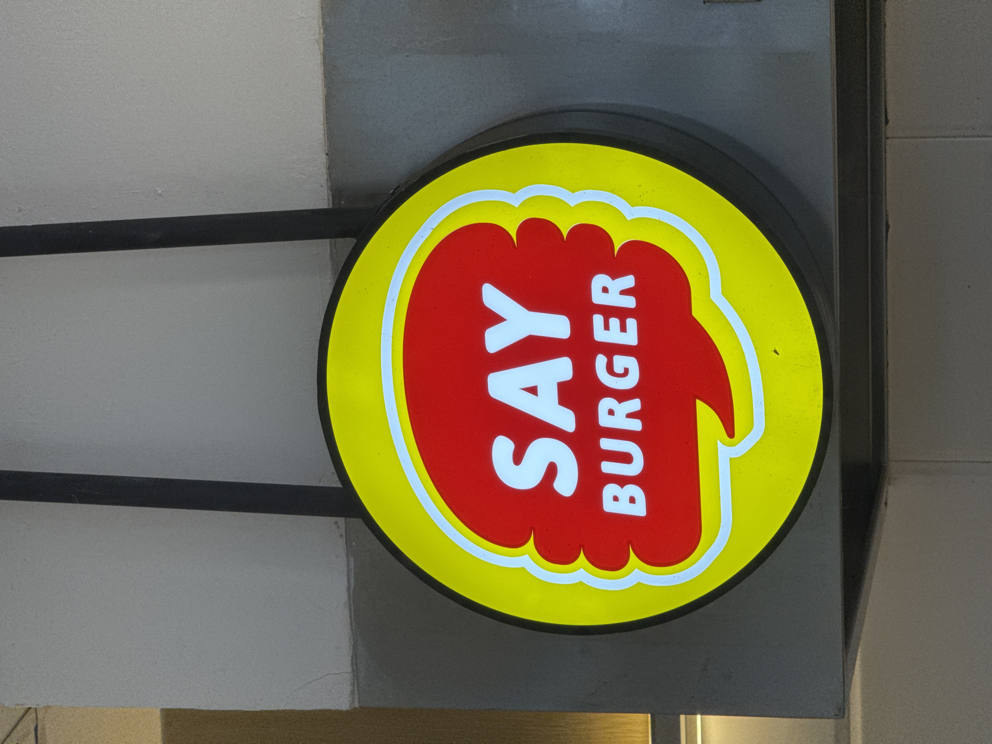 A circular, illuminated sign for a burger restaurant is the central focus, appearing to hang from an overhead structure. The sign features a bright yellow outer ring surrounding a prominent red shape that resembles a stylized burger patty or bun. A thin white outline separates the red and yellow sections.
The text "SAY BURGER" is displayed in white capital letters. "SAY" is oriented horizontally across the center of the red shape, while "BURGER" is written vertically along its right side. The sign is attached to a black metal bar, which in turn is mounted to a grey, textured ceiling or wall.
The surrounding environment consists of plain grey architectural surfaces, suggesting the sign is located under an overhang or within a commercial building in Kuta, Indonesia. The illumination of the sign, coupled with the somewhat muted lighting of the background, might indicate it is evening or night, or simply a shaded area during the day. The sign's surface shows some minor specks or imperfections.