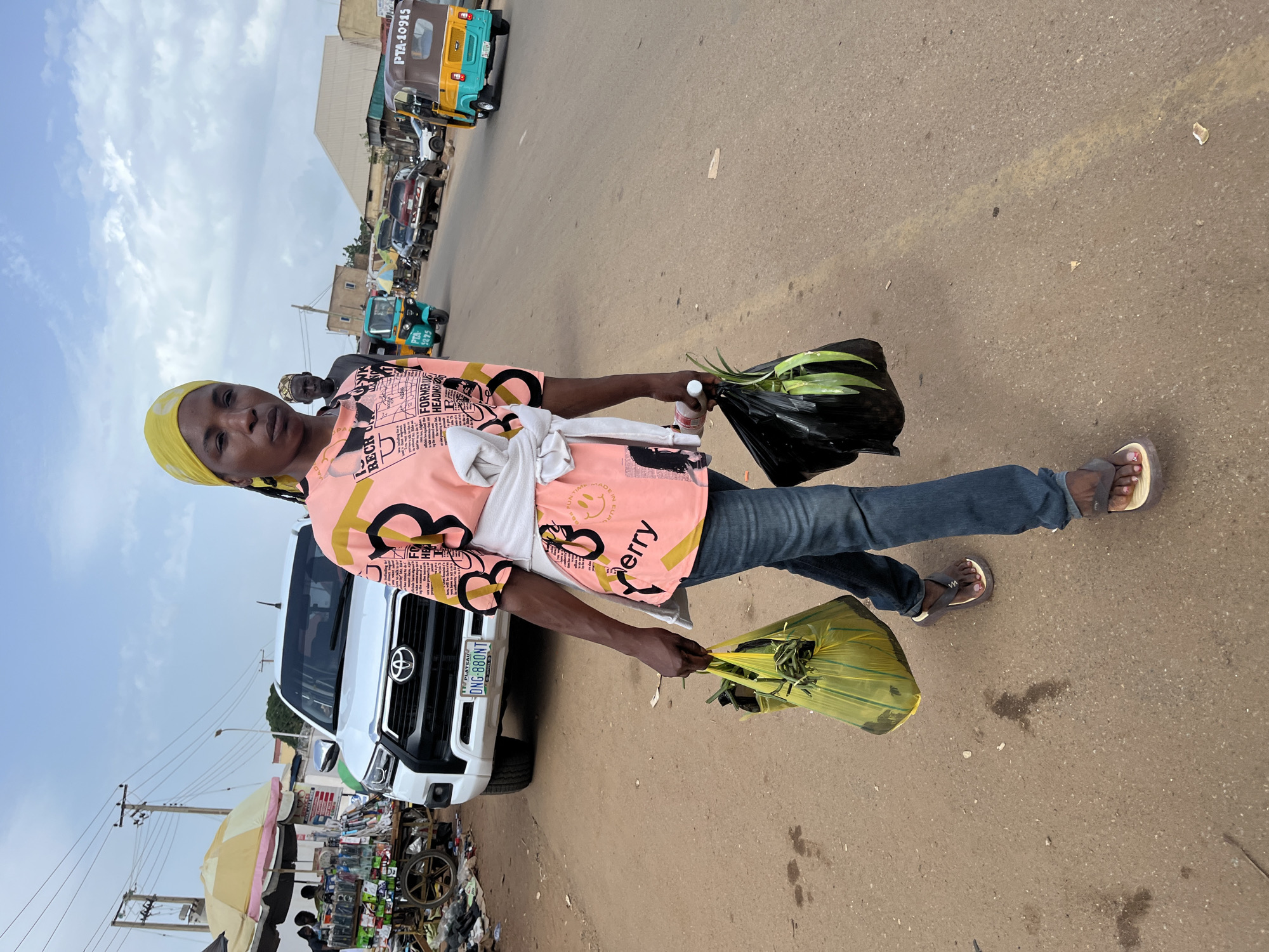 A woman wearing a yellow headscarf and a pink t-shirt with black graphics is walking on a dirt road in Jos, Nigeria. She is carrying a yellow plastic bag filled with green vegetables in her right hand and a black plastic bag containing more green vegetables in her left hand. She is wearing blue jeans and brown flip-flops. Behind her, a white SUV with a license plate reading "DNG 880NT" is parked near a market stall with umbrellas and goods displayed. Several other vehicles, including a turquoise and yellow auto-rickshaw, are visible further down the road. The sky is overcast with clouds.