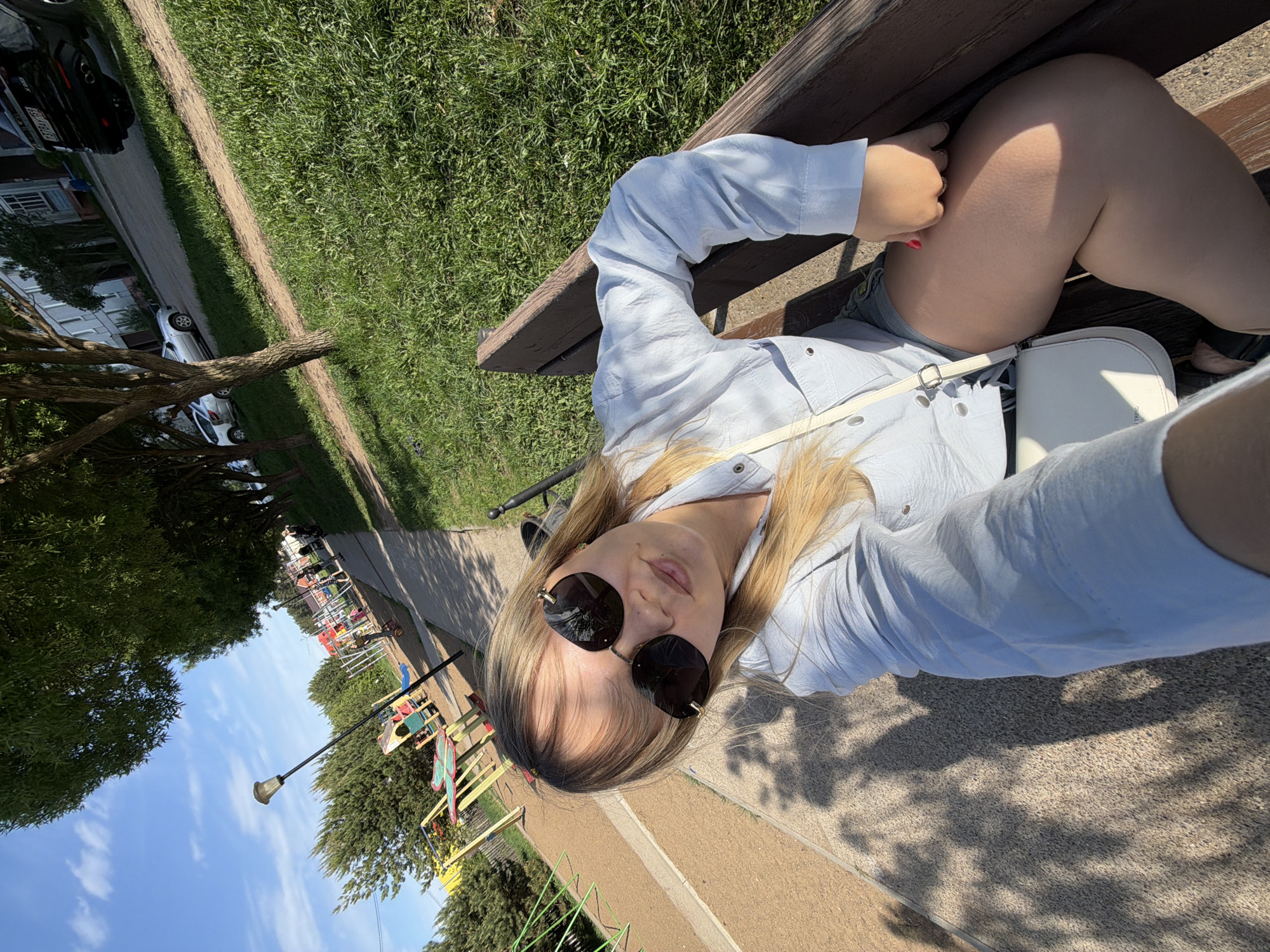 A woman is sitting on a park bench in Chaikovsky, Russia. She is wearing a light blue shirt and sunglasses. She has a white purse slung over her shoulder. In the background, there is a playground and a park.