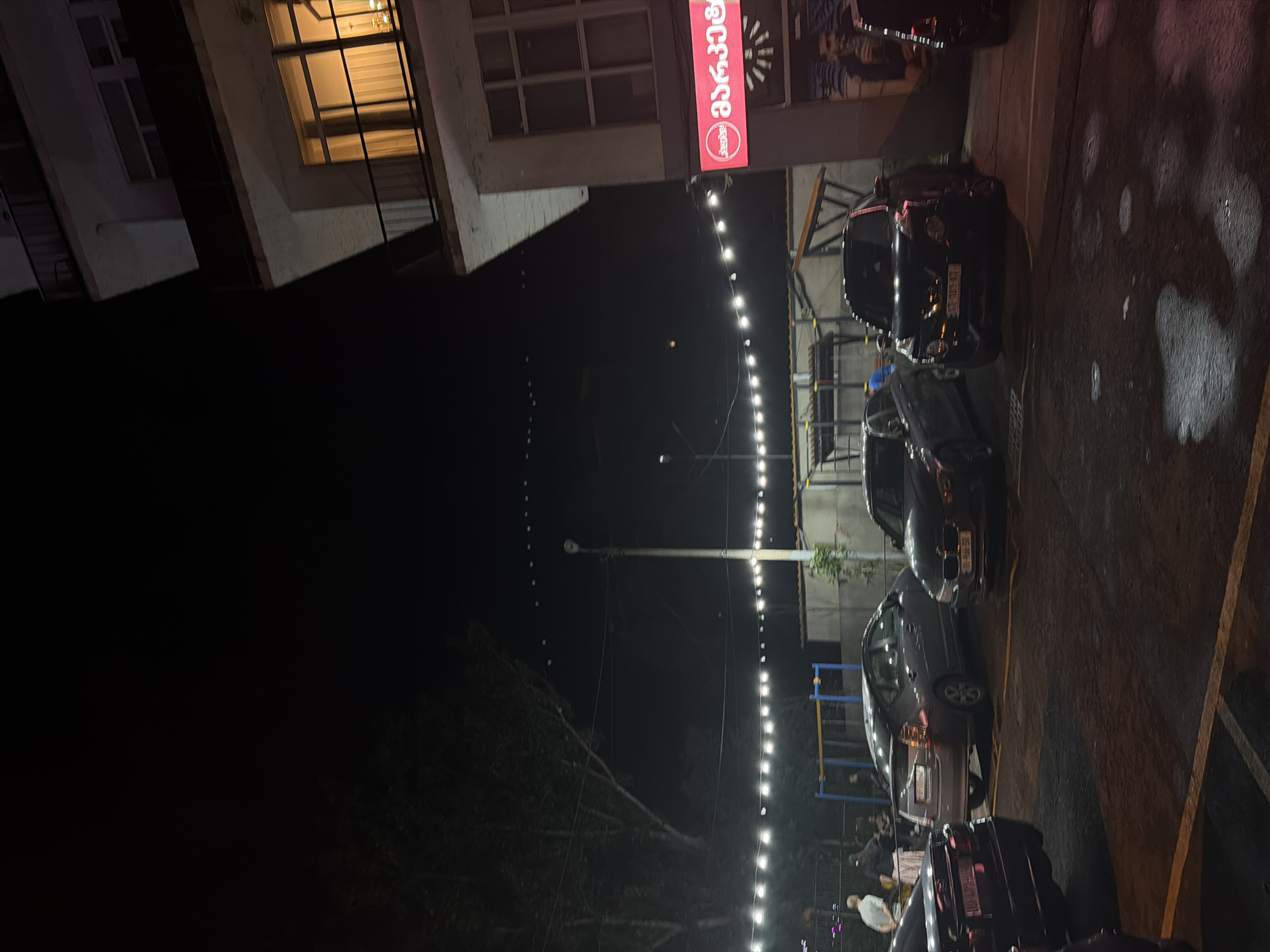 The image depicts a nighttime scene in the city of Tbilisi, Georgia, likely a parking lot area adjacent to a building. In the foreground, several cars are parked, including a dark sedan on the left, a grey hybrid sedan in the middle, and a black SUV on the right. The ground is asphalt, appearing wet in patches, reflecting the light from overhead string lights.
The building on the right is multi-story, with balconies and windows. One window on an upper floor is illuminated, casting a warm glow. A prominent red sign with white lettering is mounted on the building, featuring Georgian script and a circular logo, possibly indicating a business or establishment.
In the background, trees with dense foliage are visible on the left, silhouetted against the dark night sky. A street lamp stands tall among the trees. Further in the background, a playground structure with blue and yellow bars can be seen. There are a few individuals visible, appearing to be standing or walking, but their details are indistinct due to the low light conditions. The overall atmosphere is that of a dimly lit urban environment at night.