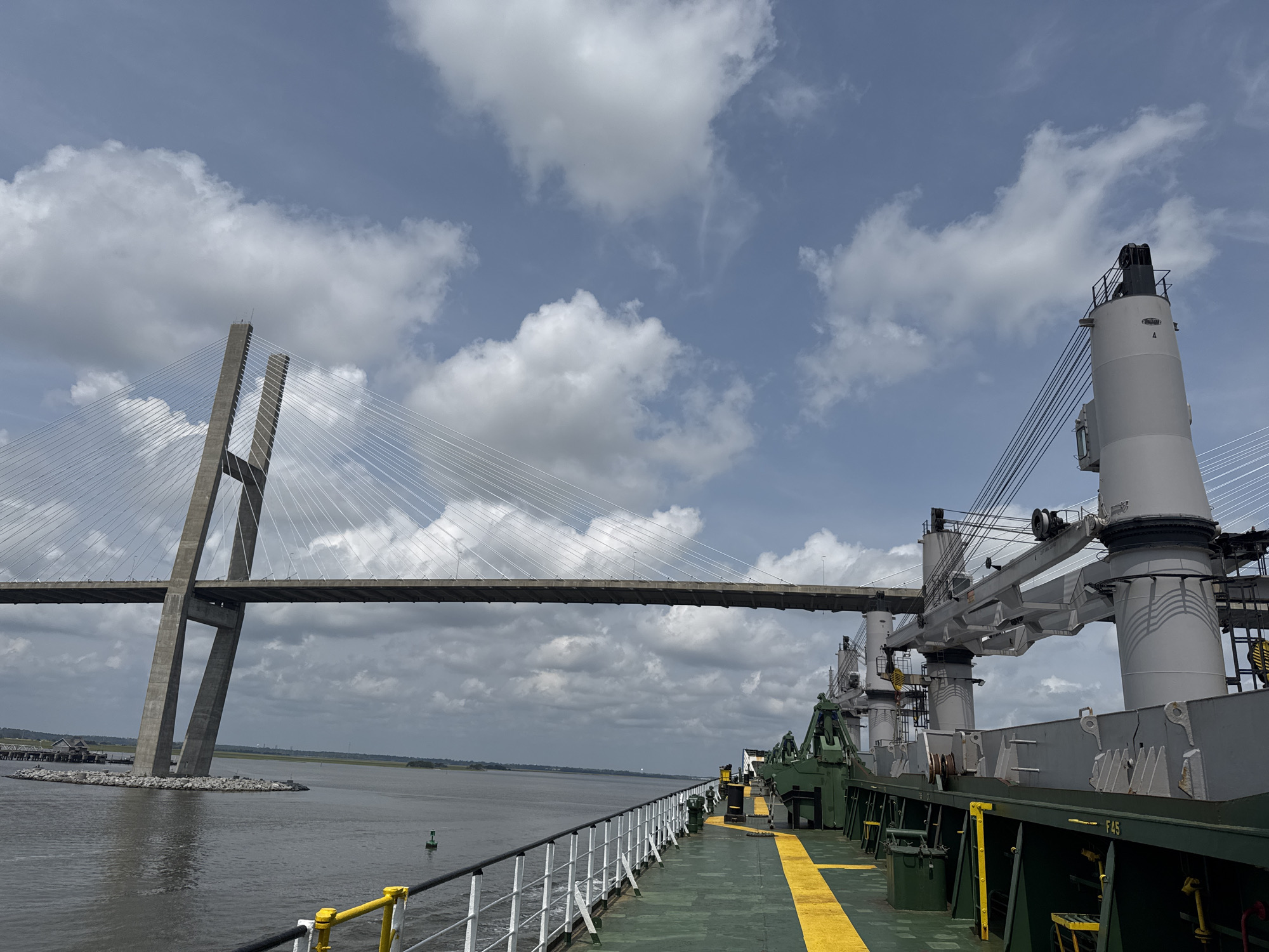 The image shows a wide shot of the St. Simons Sound near Brunswick, Georgia. The iconic Sidney Lanier Bridge, a cable-stayed bridge, spans the waterway. On the right side of the frame is the deck of a cargo ship, featuring multiple large cranes and a green hull with yellow safety lines. The water is calm and reflects the cloudy sky, which is filled with white cumulus clouds against a light blue backdrop. The bridge's concrete towers and suspension cables are clearly visible, and the ship's railing runs along the foreground. The scene depicts a busy port or shipping channel under a partly cloudy sky.