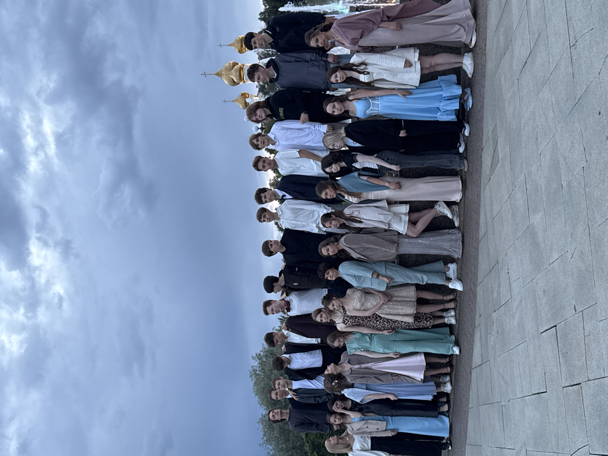This is a vertical, full-shot photograph of a large group of people, predominantly young adults, posing in two distinct lines. The individuals are dressed in formal or semi-formal attire, with the women wearing a variety of dresses and the men in suits and dress shirts. They are arranged in a staggered formation, with one line slightly behind the other, and they are all leaning backward as if posing for a group photograph. The individuals are facing the camera.
The setting appears to be outdoors, with a paved walkway or patio in the foreground and a cloudy sky in the background. There are also glimpses of what appear to be golden church domes in the upper left portion of the image, suggesting a location near a religious or historical site. The lighting suggests it is daytime, likely late afternoon or early evening, given the soft, diffused light and the overcast sky. There is no visible text in the image. The overall mood seems to be one of posed formality, perhaps for a graduation, formal event, or a group photoshoot.