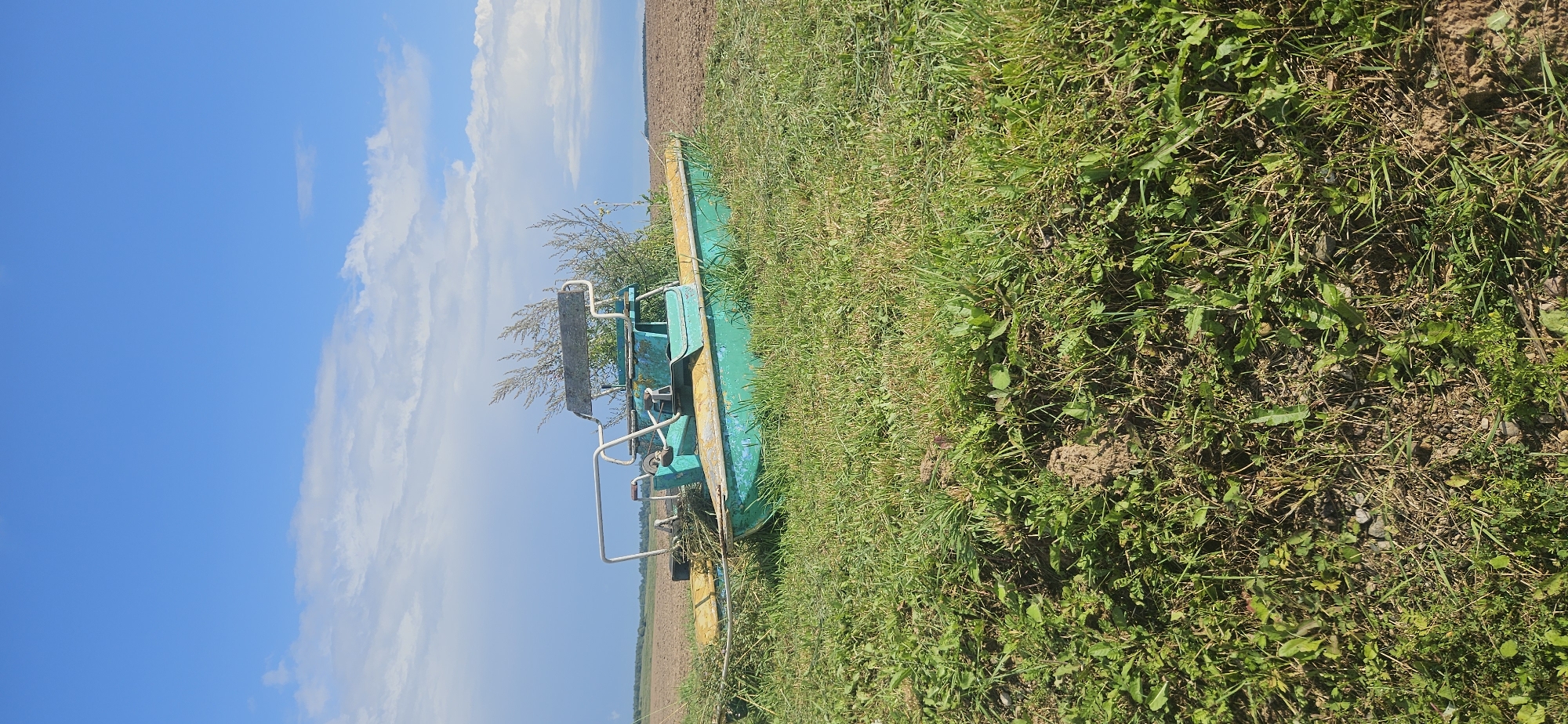 An abandoned, dilapidated green and yellow motorboat rests on a grassy embankment overlooking a tilled field under a partly cloudy blue sky. The boat is situated in a rural setting, likely in Liozna, Belarus, judging by the tilled field and the type of vegetation. The boat's hull is visibly chipped and weathered, with rust and dirt accumulated on its surface. A metal frame with seats and handlebars is attached to the boat, suggesting it was once used for recreation. Dry grass and weeds surround the boat, indicating it has been stationary for an extended period. The field beyond the embankment appears recently plowed, with rows of dark soil stretching towards the horizon. The sky above is a vibrant blue, with scattered white clouds, some of which are tinged with gray, hinting at possible distant rain. There are no people or animals visible in the scene. The overall impression is one of neglect and disuse in a pastoral landscape.