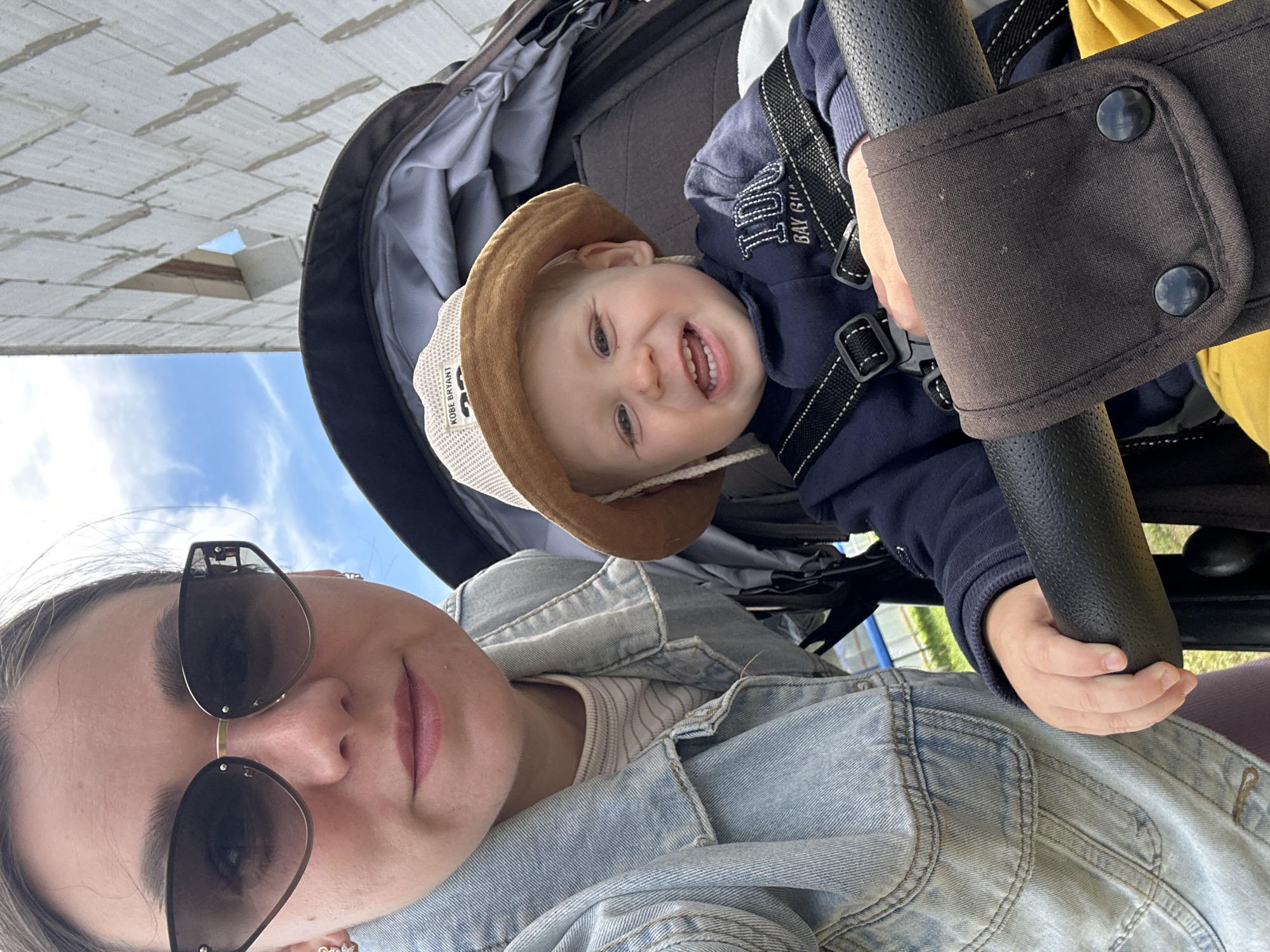 In Chaikovsky, Russia, a woman wearing a denim jacket and sunglasses is taking a selfie with a smiling baby in a stroller. The baby is wearing a sun hat and a navy blue sweatshirt. The background shows a construction site with white brick walls and a blue sky.