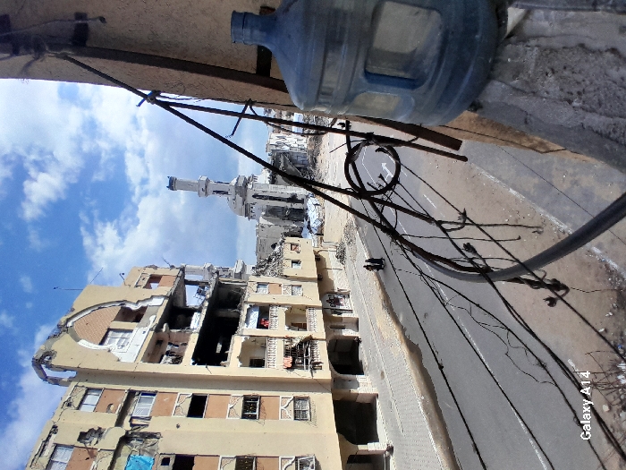 The image depicts a severely damaged urban environment in Khan Yunis, Palestinian Territory, under a partially cloudy blue sky. In the immediate right foreground, a blue plastic water bottle rests on a concrete ledge. Numerous black electrical or communication wires are strung across the right side of the frame, connected to a dark metal pole that extends vertically. On the left, a multi-story, tan-colored apartment building exhibits extensive structural damage, with large sections of its facade, balconies, and windows destroyed, exposing interior rooms, rebar, and debris; a blue tarp is visible on an upper-level balcony. Below the damaged building, a paved asphalt road extends horizontally across the mid-ground, featuring faint white directional arrow markings. Two individuals, dressed in dark clothing, walk on the left side of this road, adjacent to the damaged building. In the background, a mosque with a tall, slender minaret is prominently visible, also appearing significantly damaged, particularly its main structure. Surrounding the mosque and extending into the distance are widespread areas of rubble and collapsed buildings, indicative of extensive destruction.
