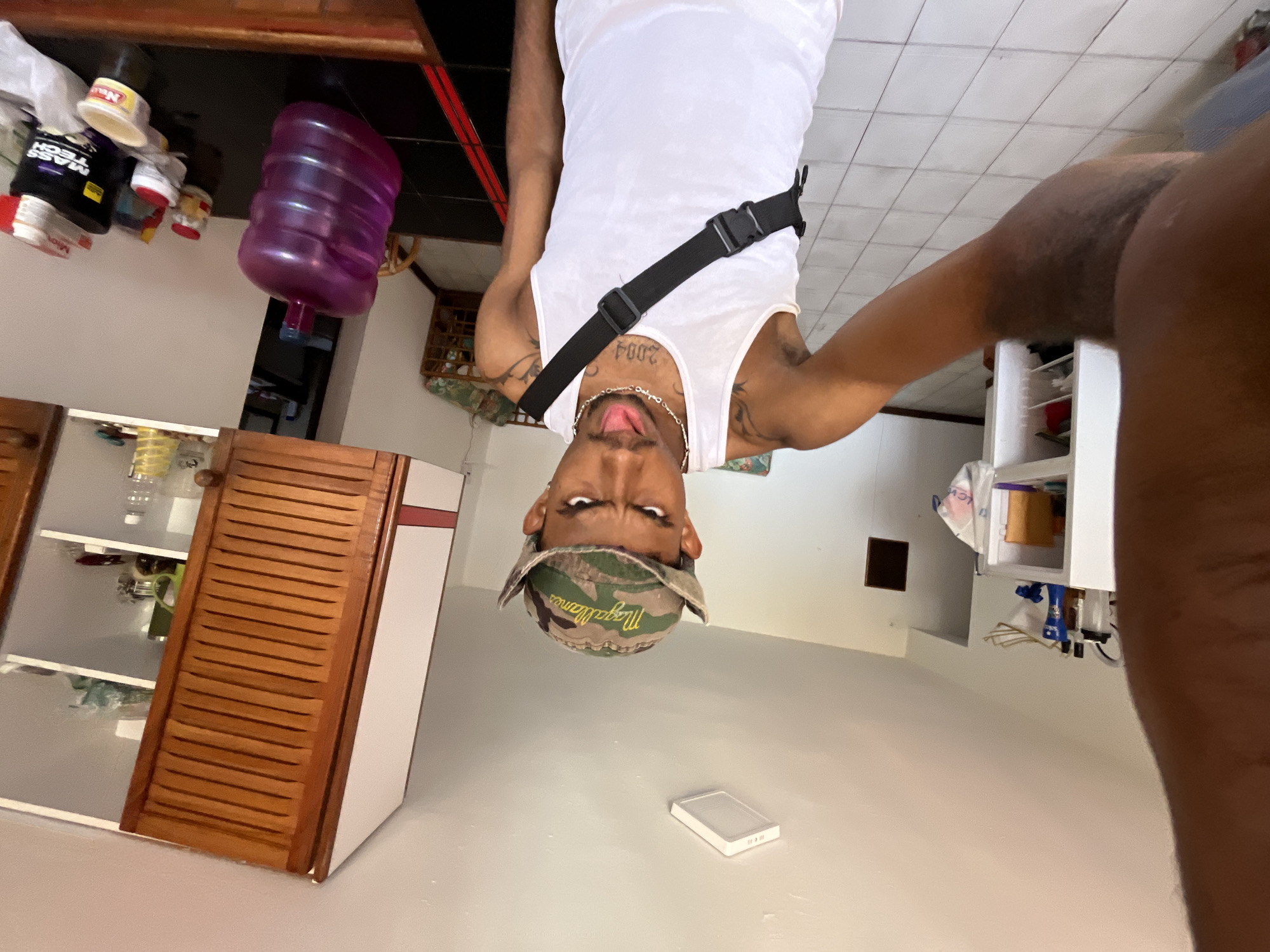 An adult male is captured in a close-up, upside-down selfie or photo, creating a disorienting perspective. He has dark skin and is wearing a white tank top, adorned with a silver chain necklace featuring a cross pendant. The number "2004" is tattooed on his chest/collarbone area, and other tattoos are visible on his arms. A black fanny pack or crossbody bag is strapped across his chest. He wears a camouflage baseball cap backward, with the word "Magallanes" visible on the brim, which is a reference to a Venezuelan baseball team. His expression is intense, with wide eyes and his tongue slightly extended.
The setting appears to be an indoor space, possibly a kitchen or dining area. In the background (which is the foreground in the image's upside-down orientation), to the viewer's left, a wooden table or countertop holds several items, including a large purple water cooler bottle, various supplement containers (one clearly marked "MASS TECH" and another "MILO"), and a white plastic bag. To the viewer's right, there are white cabinets or shelves containing miscellaneous items, and a wooden cabinet with louvered doors. The floor (or ceiling, given the image orientation) is white and tiled. A white rectangular object, possibly a smartphone or portable charger, rests on the floor. The overall lighting suggests an indoor environment. The pose and unusual orientation indicate a deliberate and possibly playful photographic moment. The "Magallanes" cap further connects the image to the provided location context of Porlamar, Venezuela.