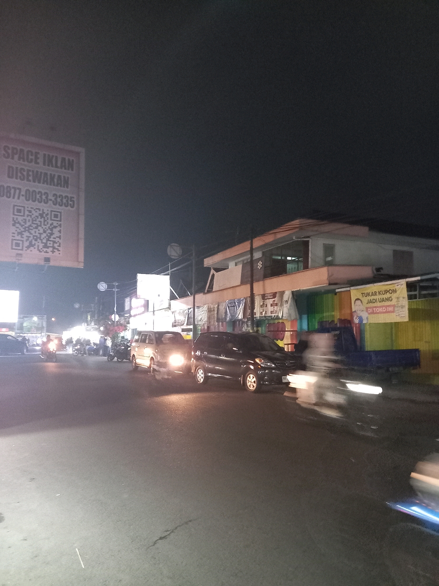 This is a nighttime photograph taken from a street level in Bawang, Indonesia. The scene depicts a moderately busy street with several vehicles, including cars and motorcycles, and a few pedestrians. The overall atmosphere is illuminated by streetlights and the headlights of vehicles.
On the left side of the image, a large, illuminated sign is visible, displaying text in Indonesian and a QR code. The text appears to advertise "SPACE IKLAN DISEWAKAN" (Advertising Space for Rent) followed by a phone number. Further down the street, several motorcycles are parked, and a few people are gathered around them, suggesting a local gathering spot or a place of business.
In the center and right of the image, cars are driving on the road. One car, a silver minivan, is stationary or moving slowly with its headlights on, casting a bright light. Another black car is visible next to it, also with its headlights on. Further to the right, a building with multiple levels is partially visible, with colorful shutters or panels on its lower section. A banner with Indonesian text, "TUKAR KUPON JADI UANG DI TOKO INI" (Exchange Coupons for Money at This Store), is displayed on the building. A blue truck is parked in front of the building.
The road surface appears to be asphalt, and there are some faint white lines marking lanes or pedestrian crossings. The sky is dark, indicating it is nighttime. The image has a grainy quality, typical of low-light photography. The presence of multiple vehicles, the advertising signs, and the building suggest an urban or semi-urban commercial area in Bawang, Indonesia. The overall impression is a snapshot of everyday life in a town at night.