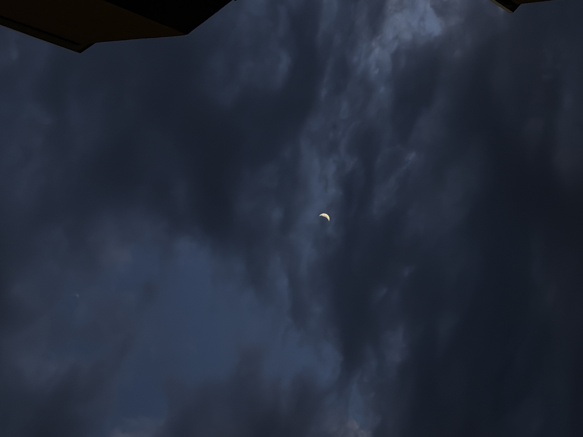 In the city of Unknown, Unknown, a thin crescent moon is visible in the dark, cloudy sky. The sky is predominantly a deep blue-gray, with thick, swirling clouds that obscure most of the celestial view. To the right of the frame, the corner of a building, likely a residence, is visible, casting a shadow into the sky. The building appears to be made of light-colored material, possibly brick or stucco, with a dark trim along its edge. The lighting suggests it is dusk or early night. There are no people or animals present in the image.