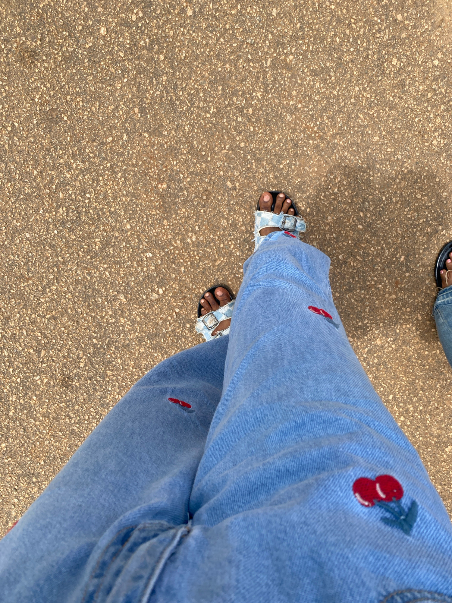 An overhead, high-angle medium shot captures a pair of legs clad in light-wash denim jeans with embroidered red cherry motifs and black-and-white checkered slide sandals, positioned on a textured, sandy-colored asphalt surface. The scene is devoid of any discernible landmarks or features that could definitively place it within the city of Iworoko, Nigeria, or any other specific urban or rural setting. The asphalt appears to be a common outdoor ground surface, consistent with a road, sidewalk, or path in many locations. The subject's lower legs and feet are the primary focus, with the background showing only the granular texture of the ground. There are no other individuals or objects visible in the frame, and the action appears to be stationary, suggesting a paused moment.