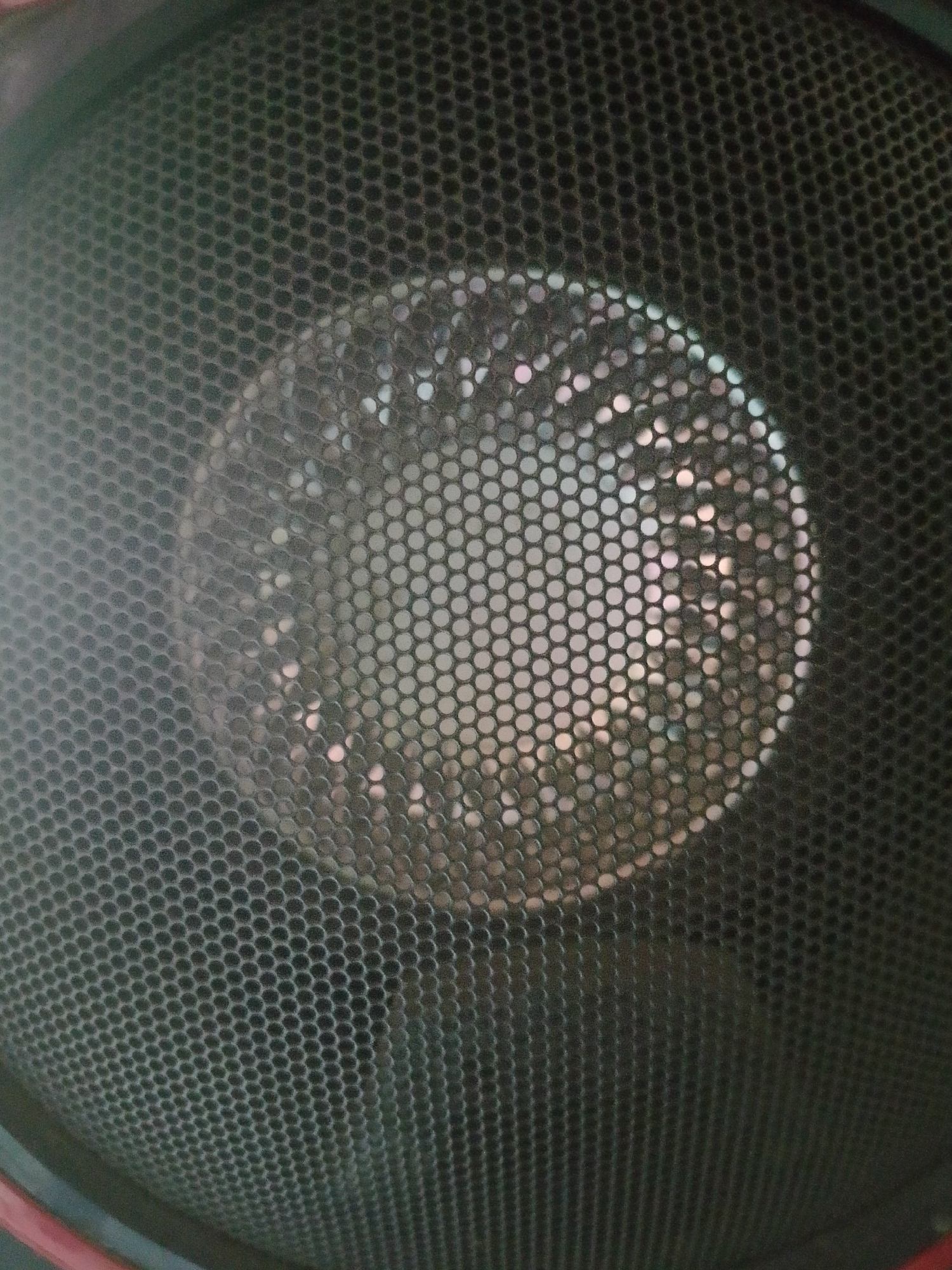 A close-up, low-angle, indoor shot shows the black mesh grille of a speaker. The grille is composed of a honeycomb pattern of small circles. In the center of the grille, a circular area of the speaker cone is visible through the mesh, reflecting light in a way that creates a blurred, shiny effect with a mix of bright white and pale pink circular highlights. The surrounding area of the speaker is out of focus. No people or specific location details are discernible.