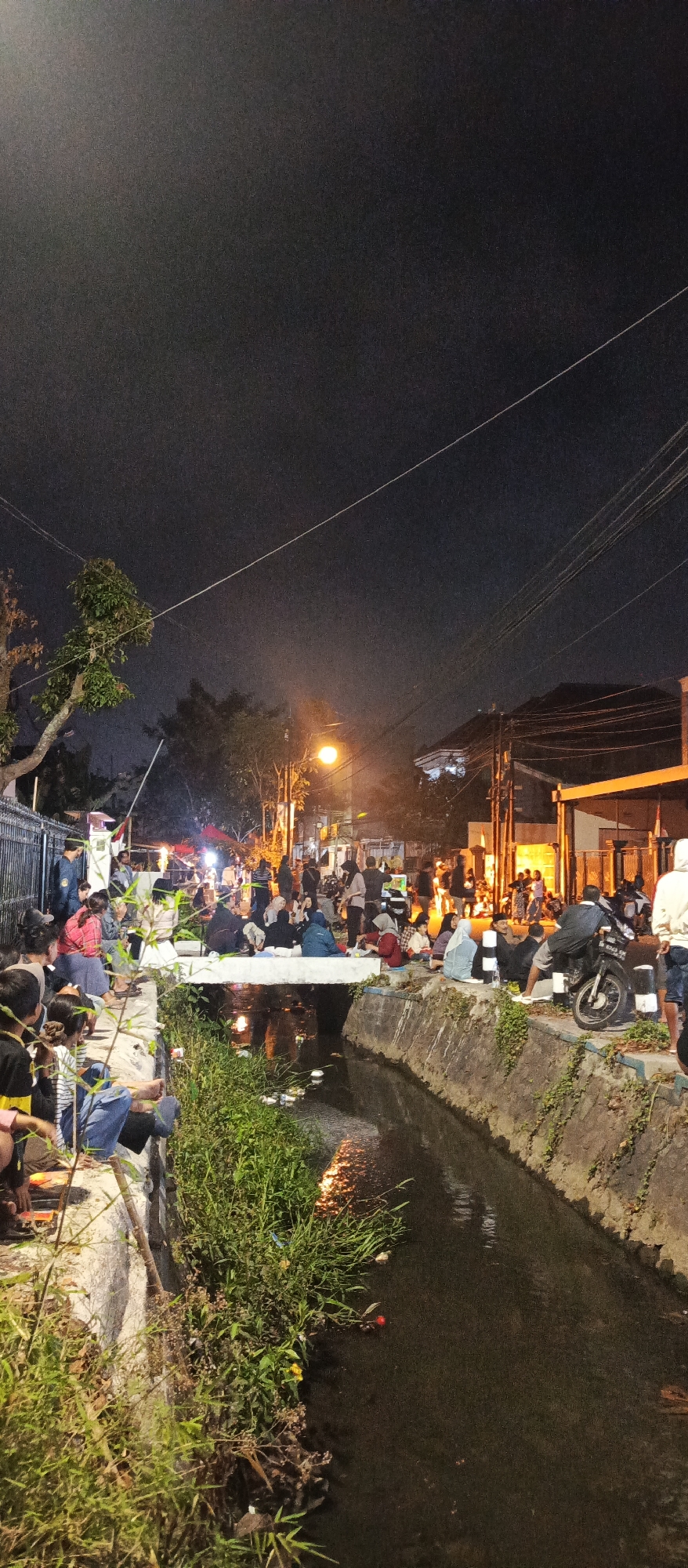 The image depicts a lively night scene in a city, potentially a street market or a gathering along a canal. Numerous people are gathered, sitting and standing on the edges of the canal and a bridge that spans it. The area is illuminated by artificial lights, including a street lamp, and there are indications of food stalls or temporary setups with lights and possibly a tent in the background. The canal runs through the scene, with water reflecting the lights and the surrounding vegetation. Several motorcycles are parked on the side of the road, suggesting a public area. The overall atmosphere is one of social gathering and activity. While the location is specified as Malang, Indonesia, this detail is not visually evident from the image itself but is provided as context.