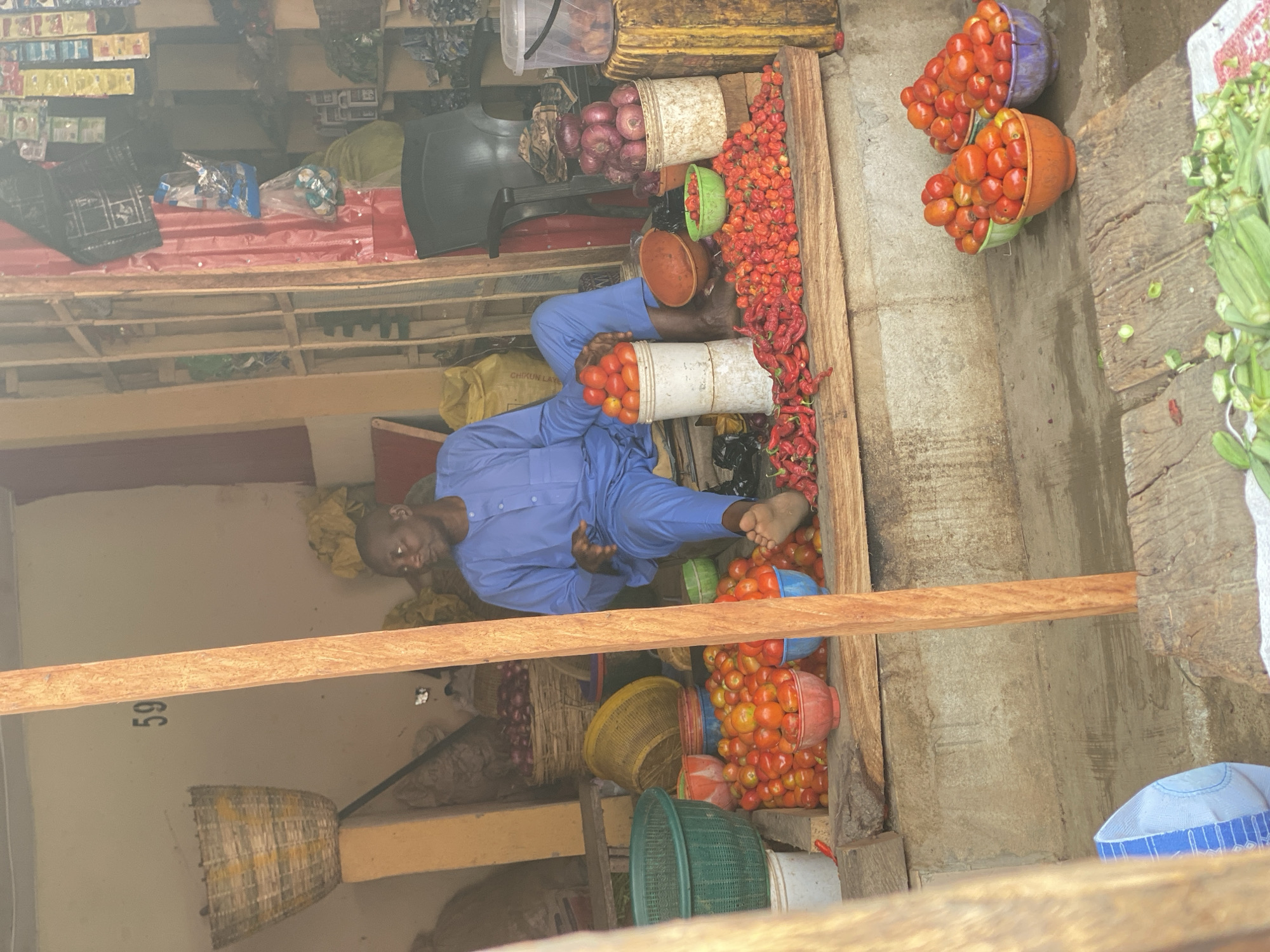 The image depicts a bustling market stall in Kafanchan B, Nigeria, seen from a slightly elevated perspective. The central subject is a man, likely a vendor, dressed in a blue traditional Nigerian outfit (a "buba and sokoto" or similar attire), sitting barefoot among his produce. He is looking towards the camera with a neutral expression, resting one arm on a white bucket filled with red tomatoes.
The stall is abundant with fresh produce, primarily bright red tomatoes overflowing from numerous colorful plastic buckets and traditional brown clay bowls. Piles of vibrant red peppers, including both small, round habanero-type peppers and longer chilies, are spread on wooden planks on the ground. Nearby, several large baskets are filled with reddish-purple onions. To the right, a pile of green pods, likely okra, is visible on a mat.
In the background, wooden shelves and racks hold various packaged goods, some wrapped in plastic. A black chair and other containers are also visible. The setting appears to be a covered market area, indicated by the wooden beams and general structure. The lighting suggests it is daytime, with some haze or filter affecting the image quality.
Visible text includes the number "59" painted on a wooden beam in the mid-ground, and partially visible text "CHIKUN LAY" on a yellow sack behind the vendor. The scene captures the essence of a local market, filled with fresh ingredients and daily commerce.
