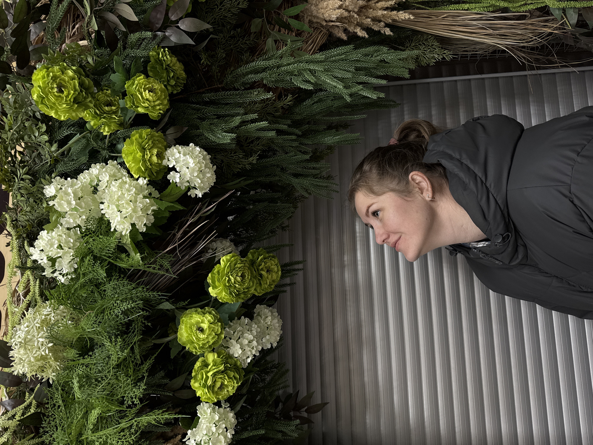 A woman in a black hooded jacket is looking at an arrangement of green and white flowers and greenery. The background appears to be a wall of vertical metal panels. The location is Горный, Russia.