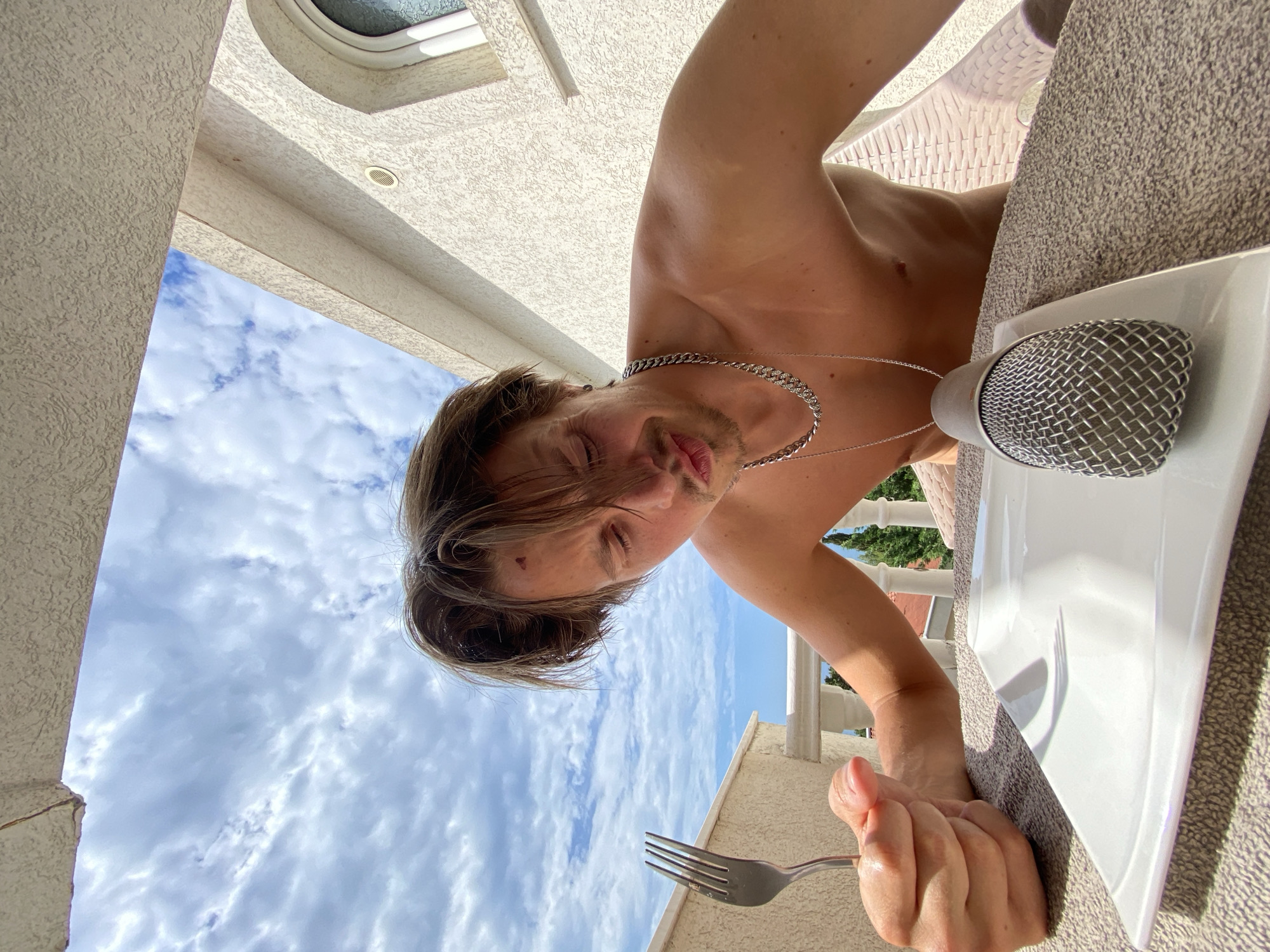 A shirtless, light-skinned male with short brown hair, a chain necklace, and a small tattoo on his forehead is photographed from a low angle against a backdrop of a partly cloudy sky. He is holding a fork in his right hand. To his right is a white plate with a microphone on it, placed on a grey patterned surface. The photo appears to be taken outdoors, near a building with textured light-colored walls. The location is Šušanj, Montenegro.