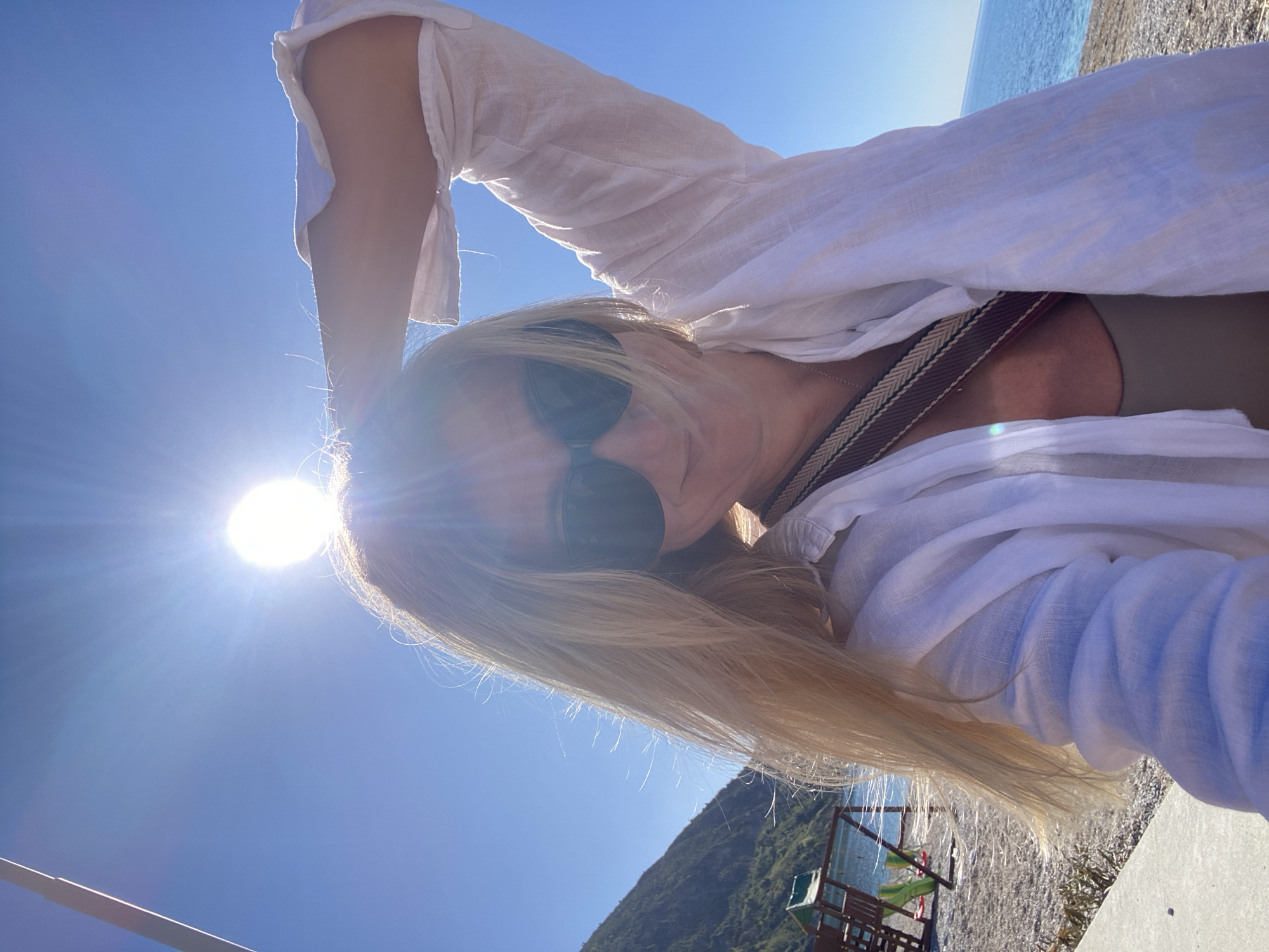 A sunlit, high-angle shot shows a woman with blonde hair wearing sunglasses and a white linen shirt. She is holding her right arm up above her head. A cross-body bag strap is visible on her chest. In the background, the Adriatic Sea meets the shoreline of Prijevor, Montenegro, with trees visible on the mountains in the distance. A lifeguard tower with chairs and tables sits on the rocky beach.