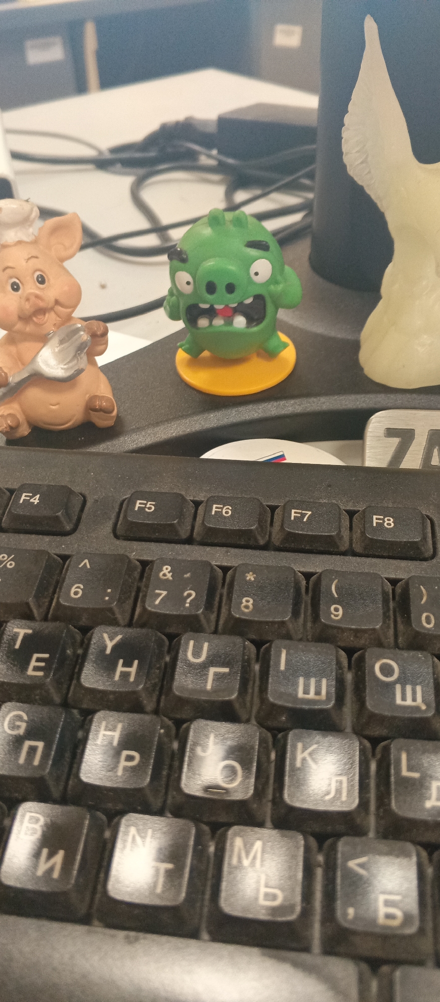 This is a close-up, high-angle shot of a keyboard with toys placed on top of it. In the foreground, a black keyboard is visible with various keys, many displaying Cyrillic characters. The keys are dusty, indicating the keyboard is in use. Above the keyboard, two small figurines are prominent. On the left is a light brown pig figurine wearing a chef's hat and holding a utensil. To its right is a green Angry Bird-like character, with its mouth open and arms raised in a defiant pose, standing on a yellow base. In the background, other objects and a blurred office-like setting are visible, including what appears to be another figurine on the far right, possibly a bird or angel, made of a light, translucent material. The overall impression is a casual office desk setup with decorative toys. No specific time of day or weather is discernible. The visible text includes key labels on the keyboard such as "F4", "F5", "F6", "F7", "F8", along with numbers and symbols, and Cyrillic letters. There is also a partial silver label with the number "7" and another partially visible label with Russian flag colors.