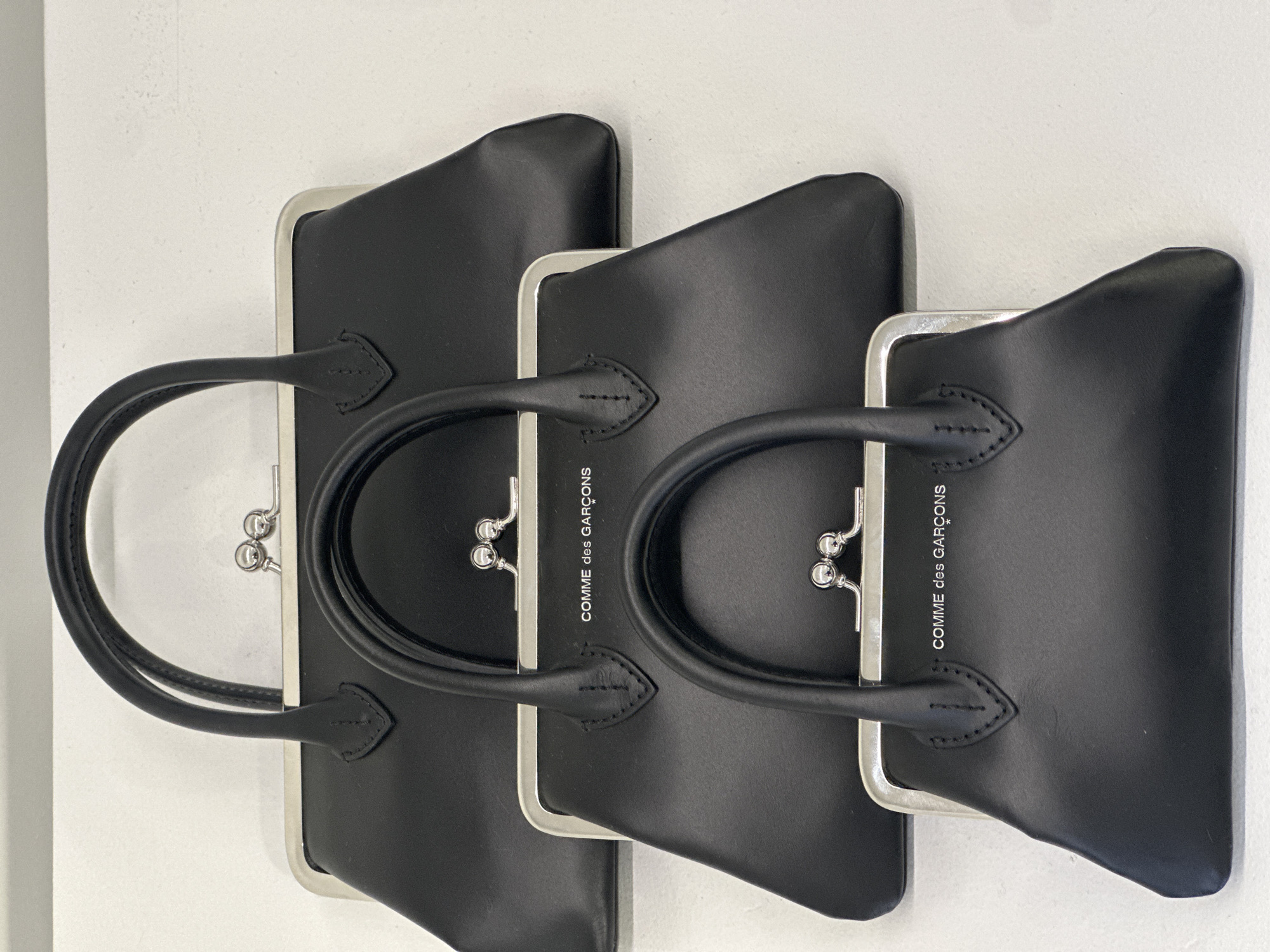 This media file displays three identical black handbags, arranged horizontally and slightly overlapping, against a plain light-colored (likely white or off-white) background. The handbags appear to be lying on their sides, with their handles extending towards the left side of the frame.
Each handbag features a structured, boxy shape with a smooth, matte finish, indicative of leather or a similar material. They are equipped with silver-toned metal frames at the top, incorporating a traditional kiss-lock clasp mechanism. Two rounded top handles, matching the black material of the bags, are attached to the frame. The handles are stitched in place, showing visible black thread details.
The bags are presented in what appears to be a clean, well-lit indoor setting, possibly a studio, with soft and even lighting that highlights their contours without harsh shadows. No specific activity is taking place; it is a static product shot.
The brand name "COMME des GARÇONS" is visibly printed in white capital letters on the side of at least two of the handbags, indicating their designer origin. The bags appear new and in pristine condition.