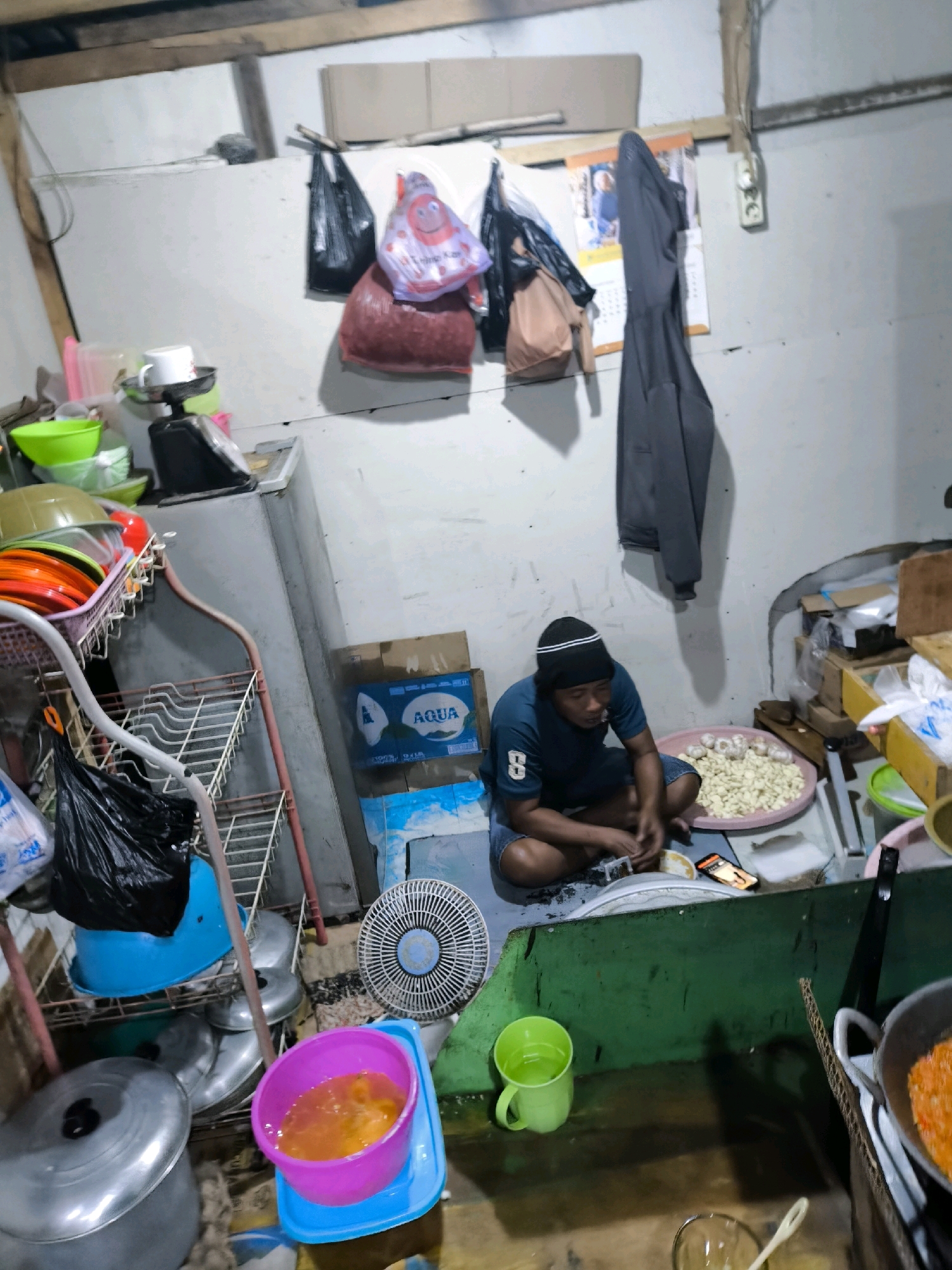 In Moluo, Indonesia, a man sits on the floor of a dimly lit room, sorting through a large pile of peeled garlic. He is wearing a dark blue t-shirt, shorts, and a black beanie. In front of him is a round, pink tray filled with garlic cloves and a few whole bulbs. To his right, a smartphone is propped up, displaying a screen. The room appears to be a kitchen or food preparation area, with various items scattered around. To the left of the man, a shelf unit holds plates, bowls, and a black plastic bag. A large refrigerator stands against the wall, and above it, several bags and a calendar hang. A small fan is positioned on the floor near the man. Further to the right, a green partition separates this area from another part of the room where a pot is on a stove, with some orange food visible inside. The walls are painted white, and the floor is concrete. The overall impression is of a functional, albeit cluttered, domestic workspace.