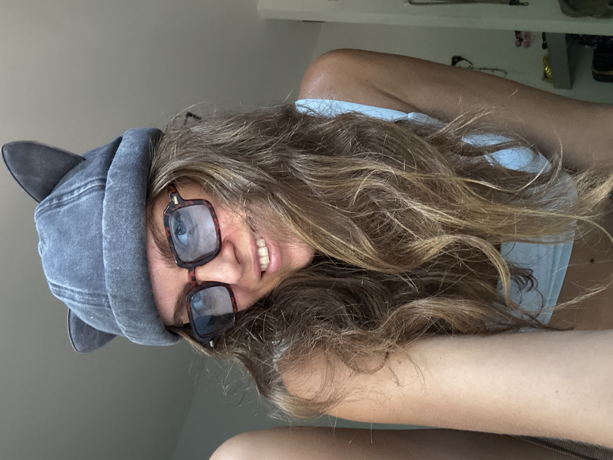 A woman with shoulder-length, wavy, brown hair with blonde highlights, wearing a gray hat with small ears, and oversized, square-framed glasses is smiling at the camera. She is wearing a light blue top. The background is blurry, suggesting an indoor setting. There is no indication of the location being in Moo 4, Thailand.