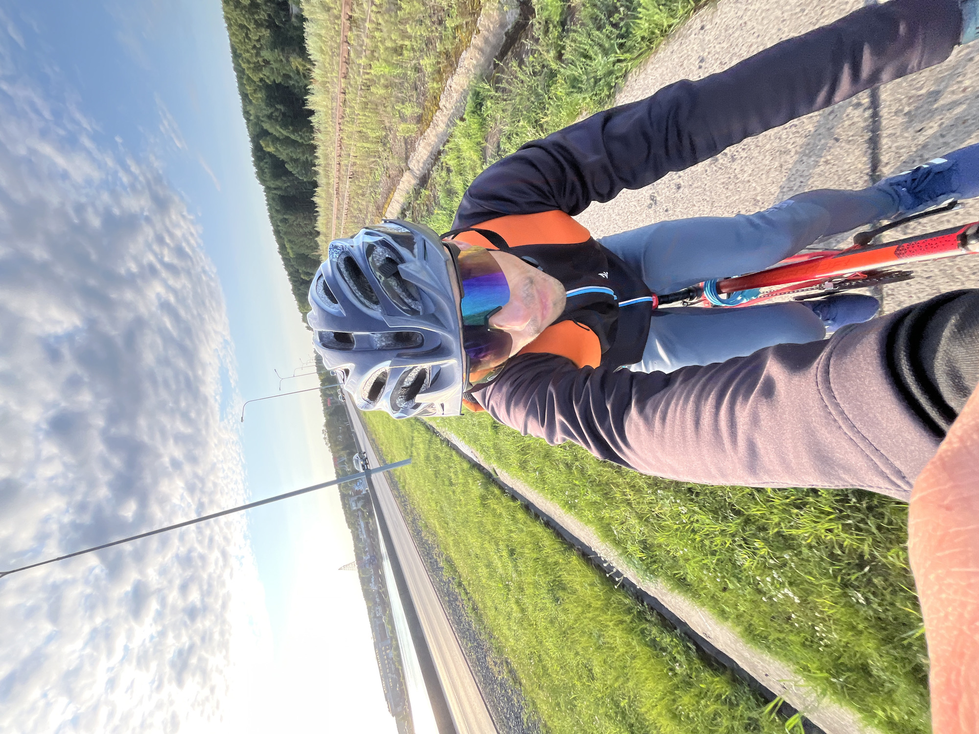 A first-person perspective selfie shows a person cycling on a paved path adjacent to a road and a body of water. The cyclist wears a dark jacket with orange accents, blue cycling pants, blue athletic shoes, a gray helmet, and reflective sunglasses. The road appears to be in the city of Chaikovsky, Russia, with some buildings and industrial structures visible in the distance on the left. The right side of the frame shows a field with some tracks running through it and a dense line of trees bordering the horizon. The sky is partly cloudy with the sun setting or rising, casting a warm light on the scene.
