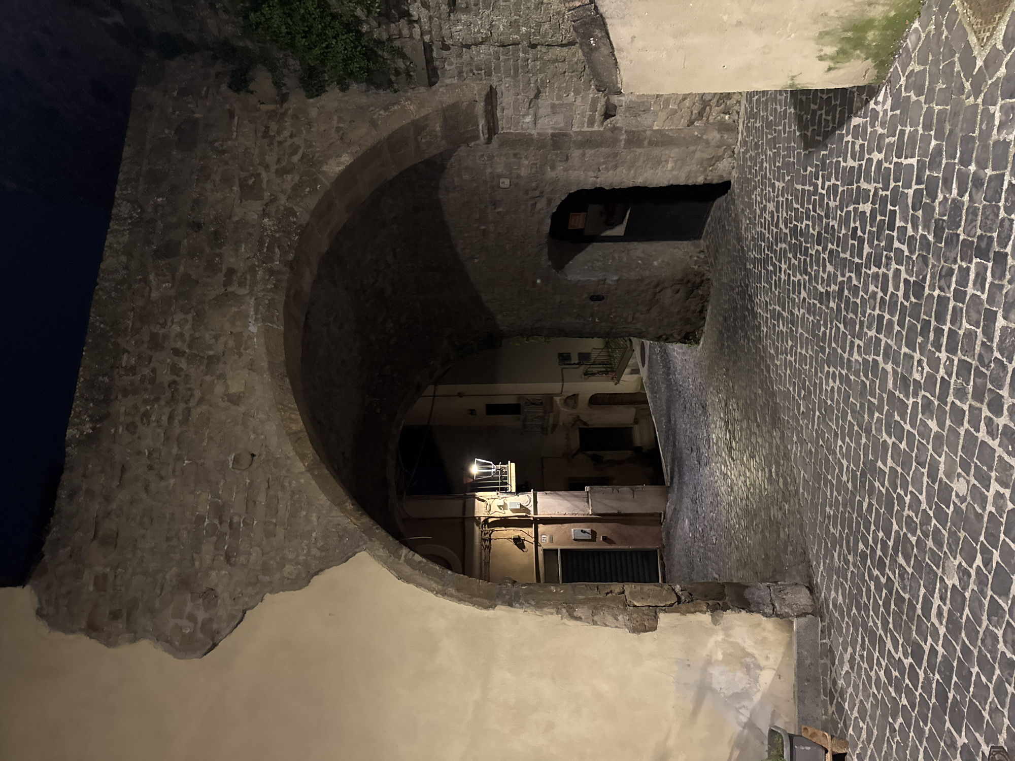 The image shows a narrow passageway in the city of Castelnuovo di Porto, Italy. The passageway is formed by an arched stone structure on the left, leading to a street paved with cobblestones on the right. A building with a lamp is visible through the passageway. The lighting suggests it is nighttime. The overall impression is of a historic or old town environment.
