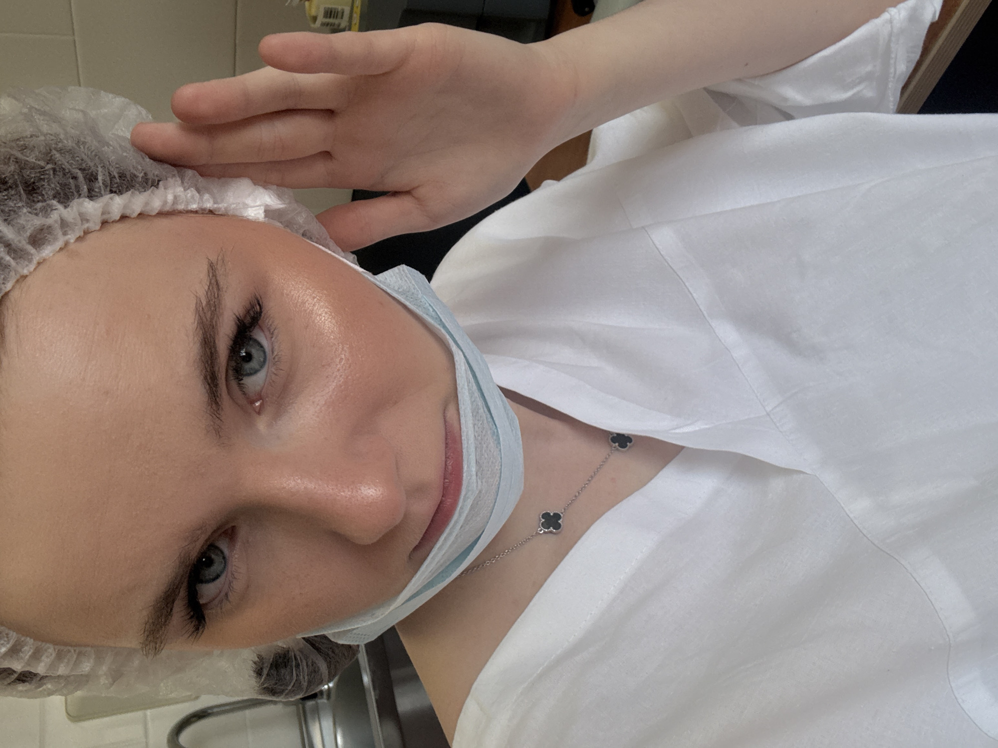 A person wearing a white bouffant cap and a surgical mask is looking at the camera, with their right hand raised near their head. They are wearing a white shirt and a silver necklace with black clover-shaped charms. The background is blurred, but shows a plain wall and a stainless steel fixture, possibly in Saint Petersburg, Russia.